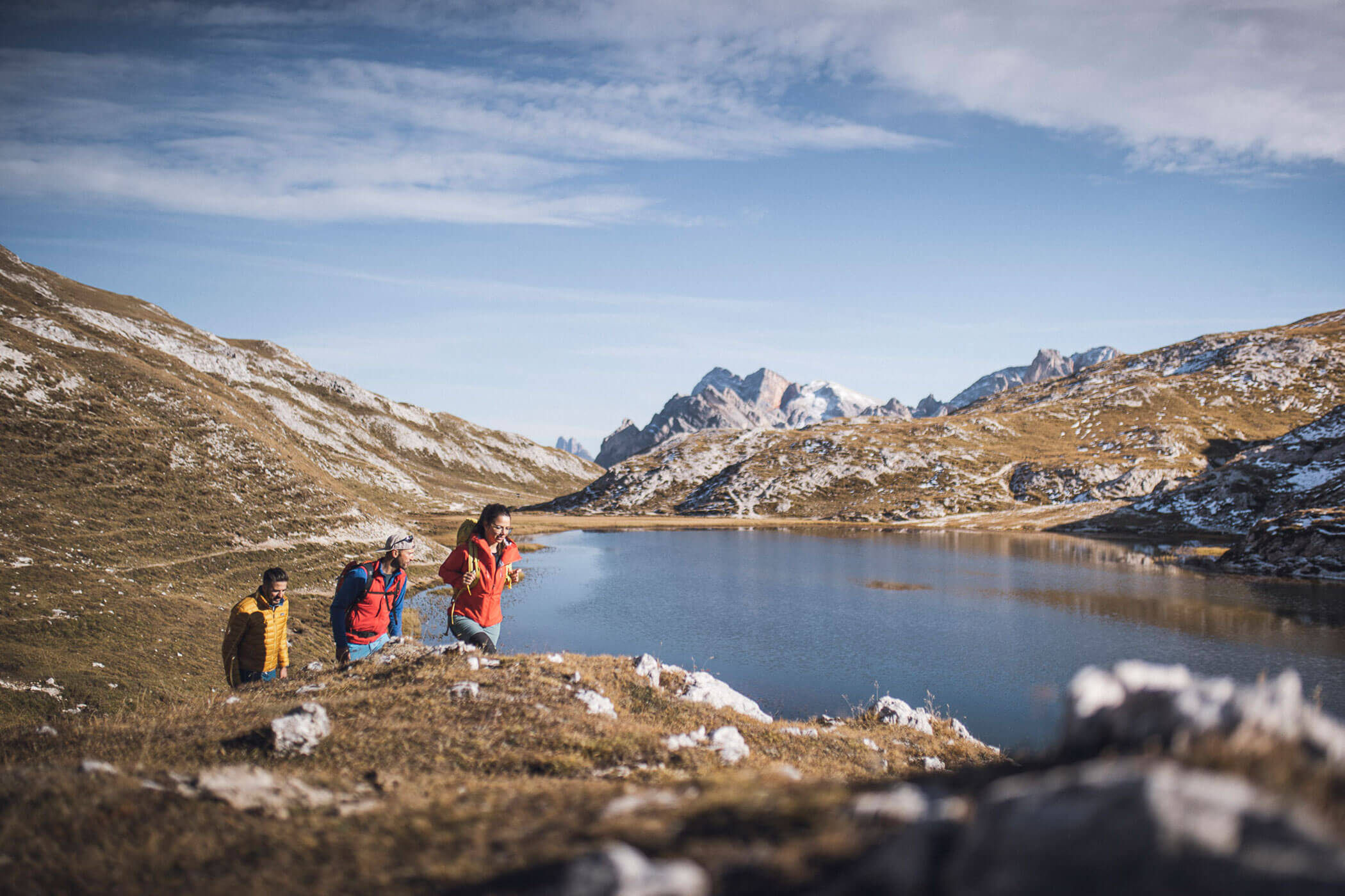 Drei Wanderer spazieren an einem Bergsee unter blauem Himmel, umgeben von felsigen Hügeln und fernen Gipfeln. - Hotel Fameli