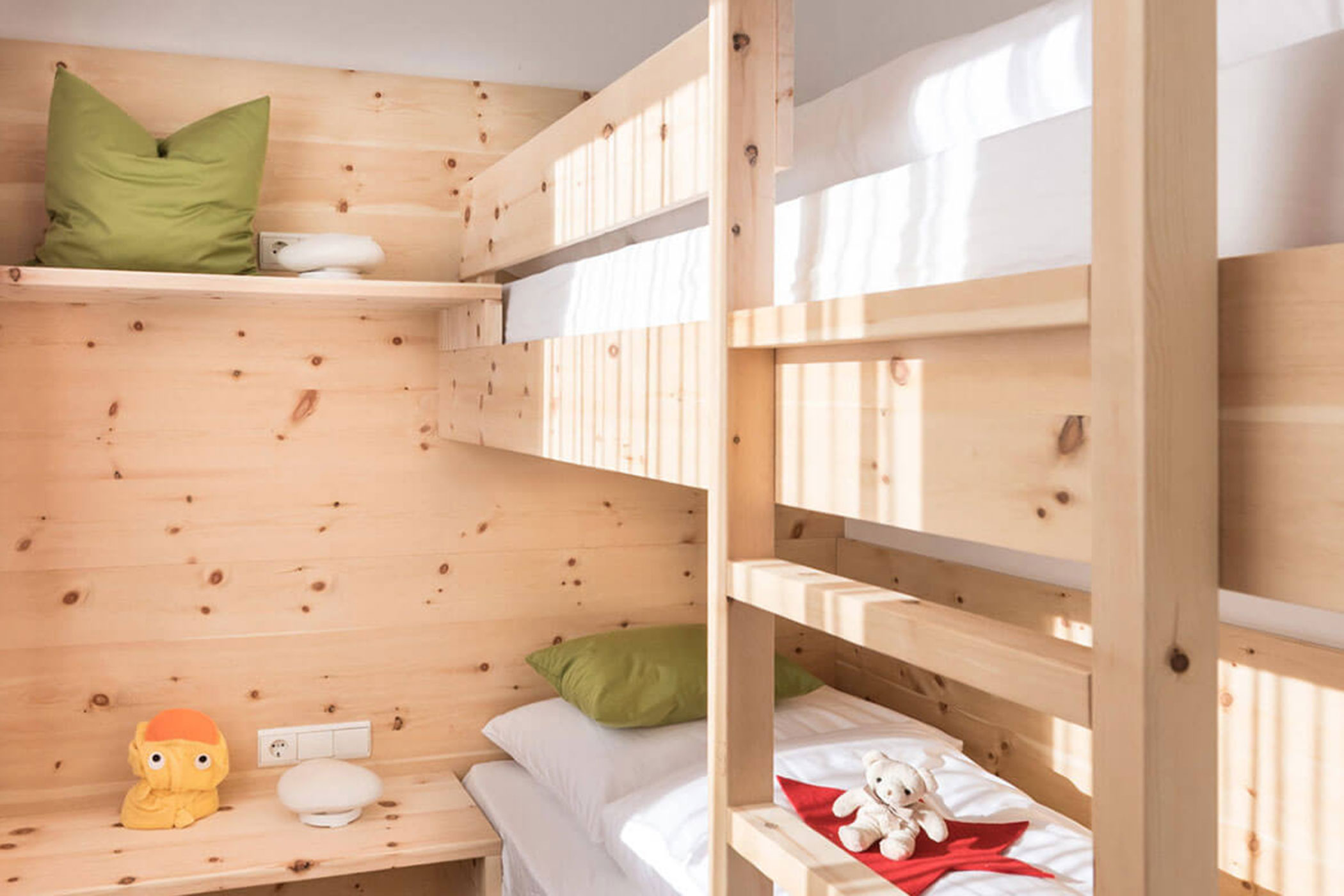 Etagenbetten aus Holz mit grüner und weißer Bettwäsche, Plüschtiere und Sonnenlicht, das in ein gemütliches Zimmer fällt. - Hotel Fameli