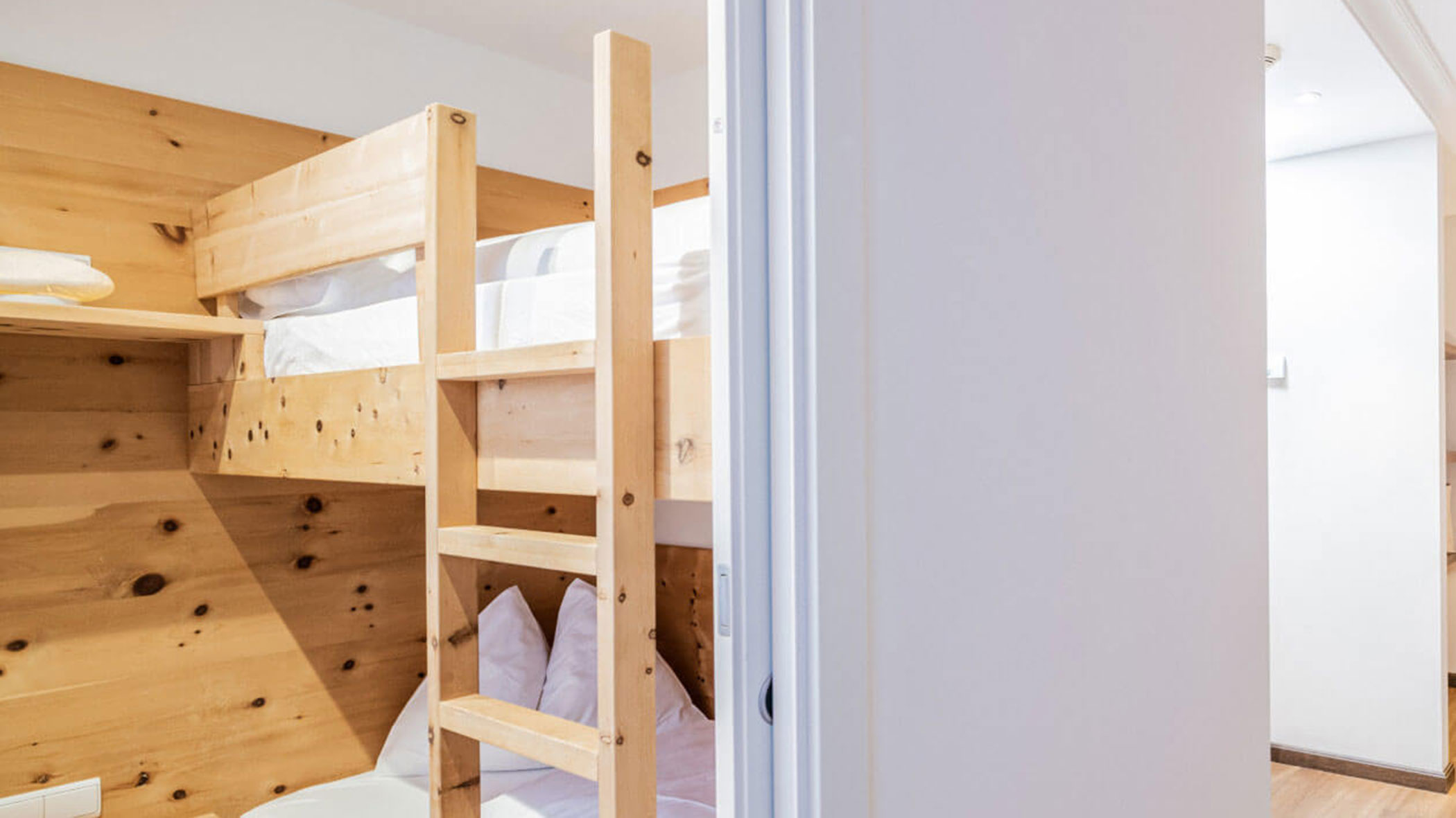 Etagenbetten aus Holz mit weißem Bettzeug in einem gemütlichen, hellen Zimmer mit teilweiser Aussicht auf einen Flur. - Hotel Fameli