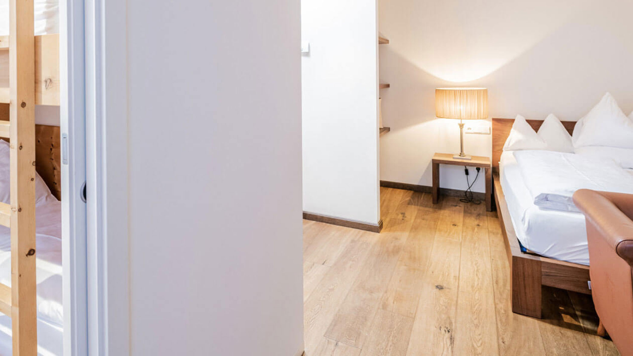 Ein helles Schlafzimmer mit Holzboden, Doppelbett, Beistelltisch mit Lampe und einem teilweise sichtbaren Etagenbett. - Hotel Fameli