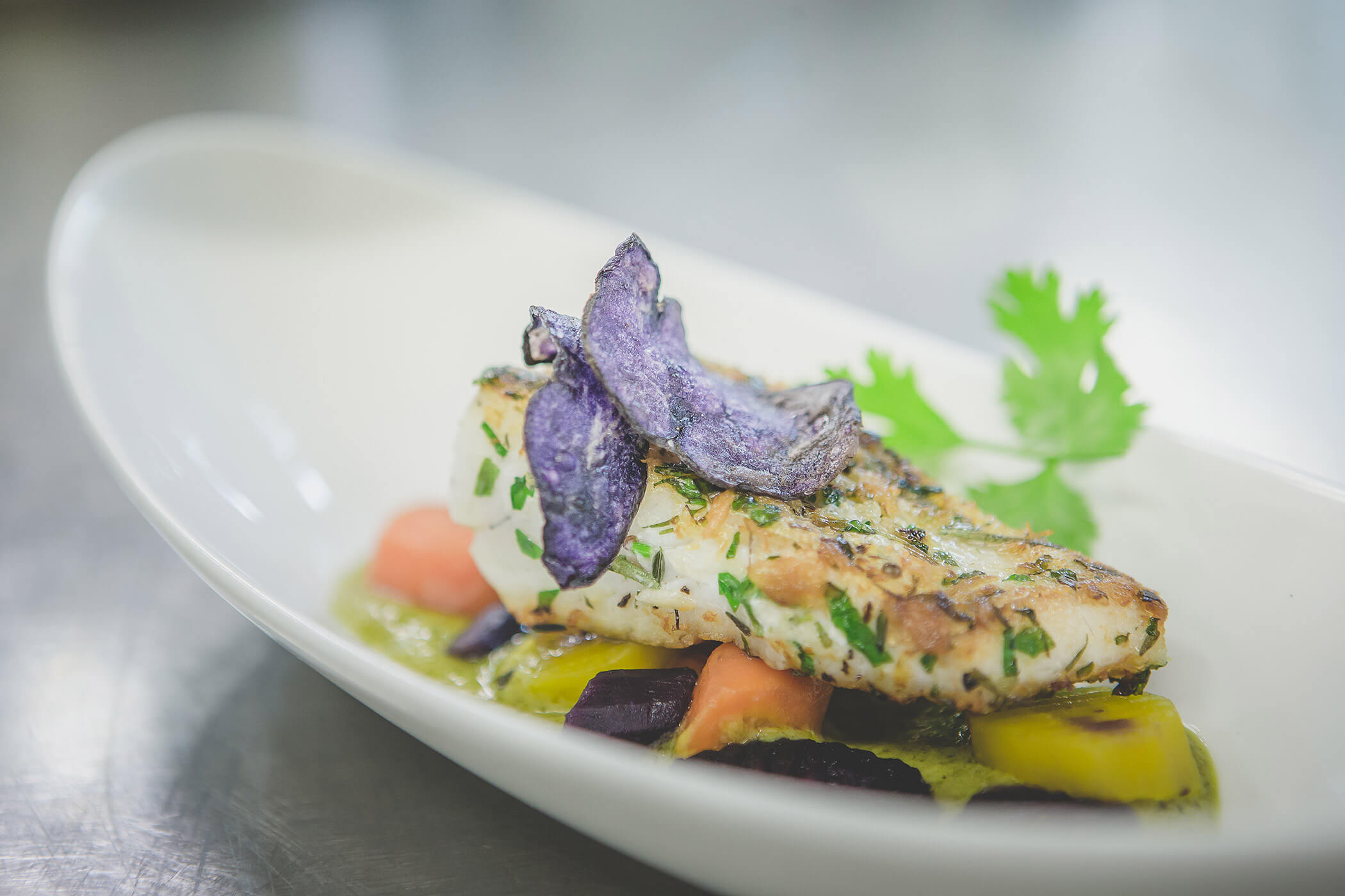 Gegrilltes Fischfilet garniert mit violetten Kartoffelchips, Kräutern und Gemüse auf einem weißen Teller. - Hotel Fameli