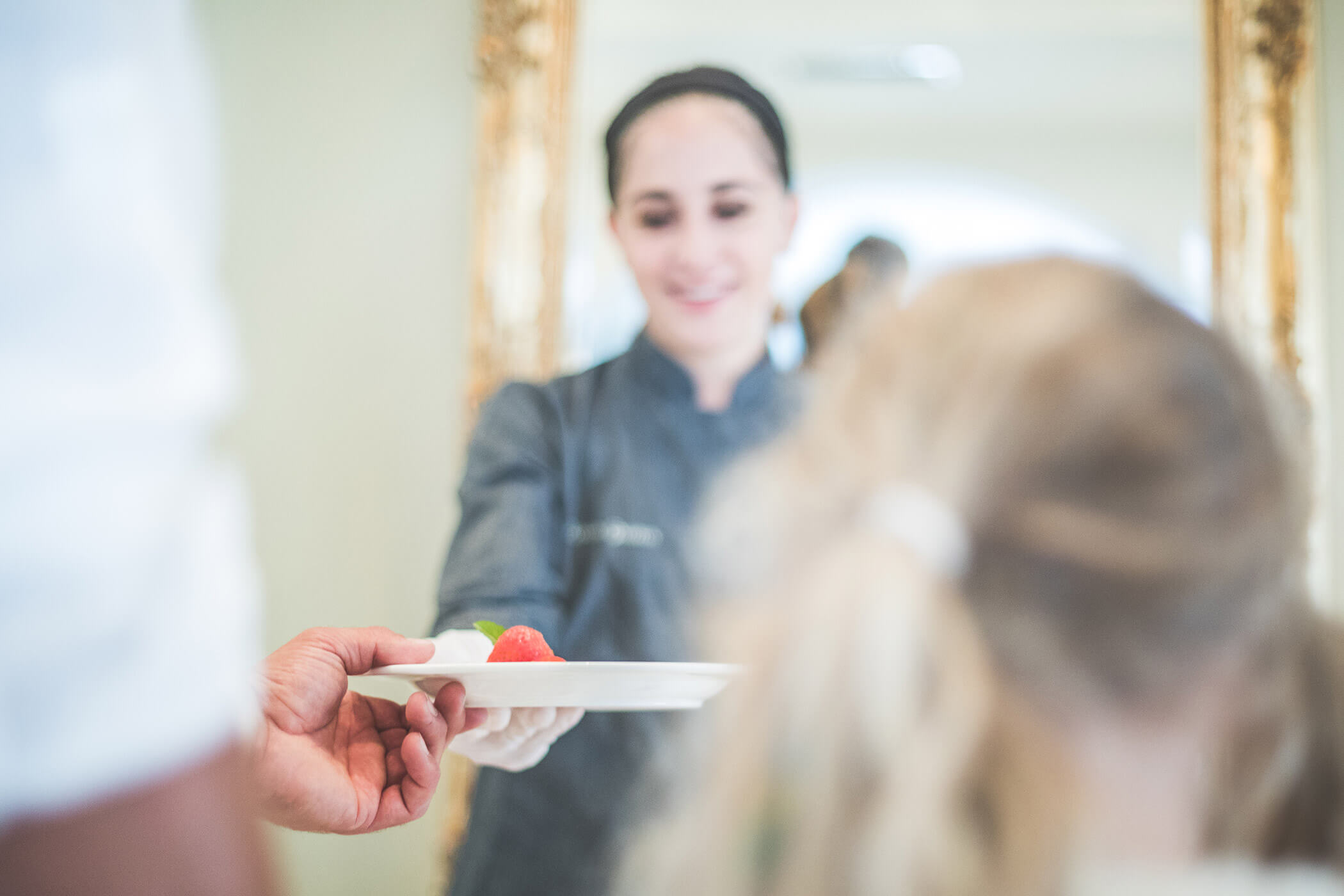 Ein Koch reicht einer Person einen Teller mit Essen, während ein Kind mit blonden Zöpfen zuschaut. - Hotel Fameli
