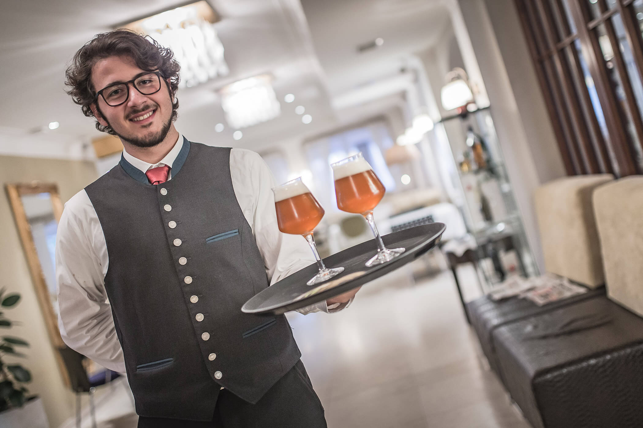 Ein lächelnder Kellner in einer Weste hält ein Tablett mit zwei Gläsern Bier in einem stilvollen Restaurant. - Hotel Fameli