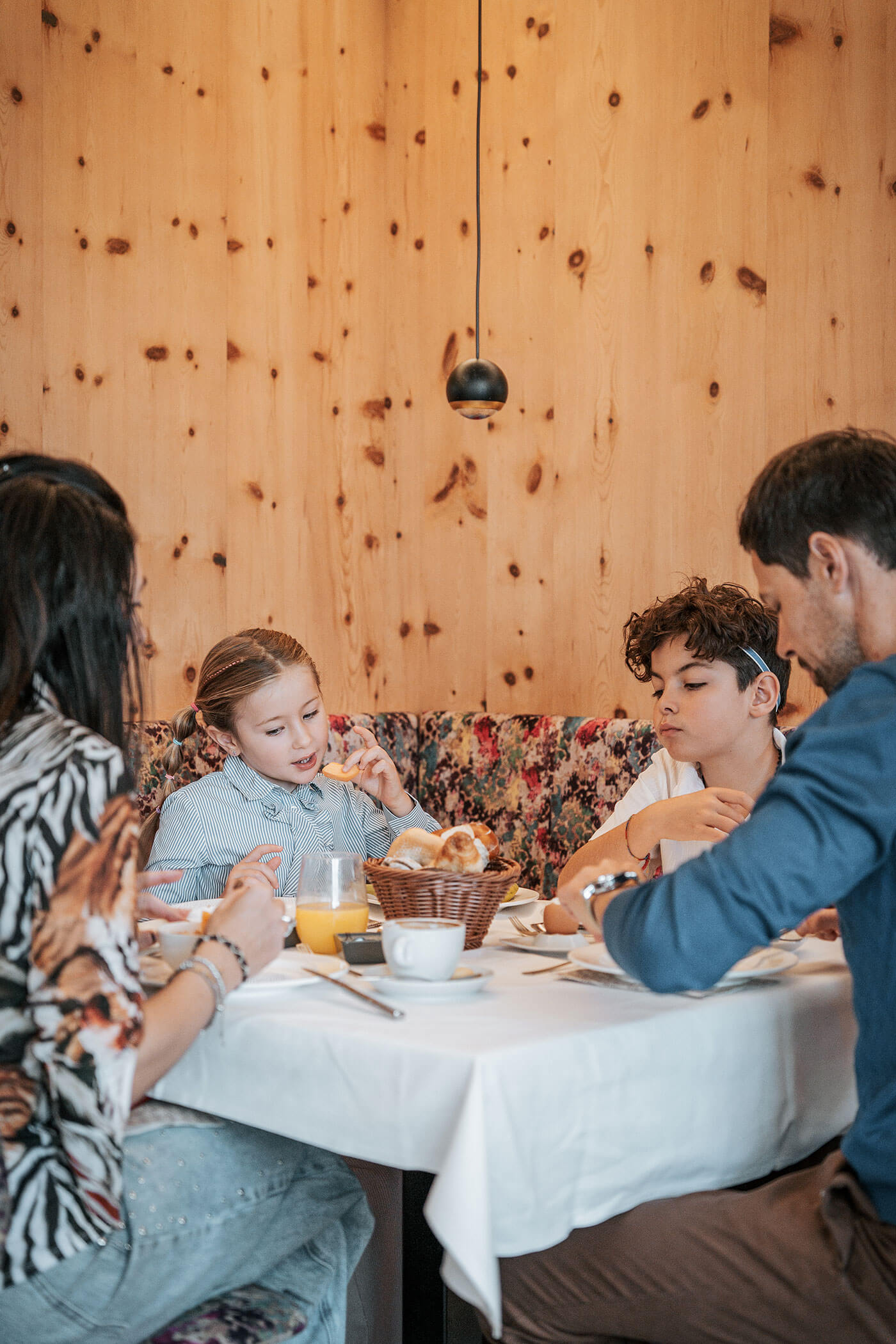 Eine vierköpfige Familie sitzt in einem gemütlichen, holzgetäfelten Restaurant am Frühstückstisch, isst und unterhält sich. - Hotel Fameli