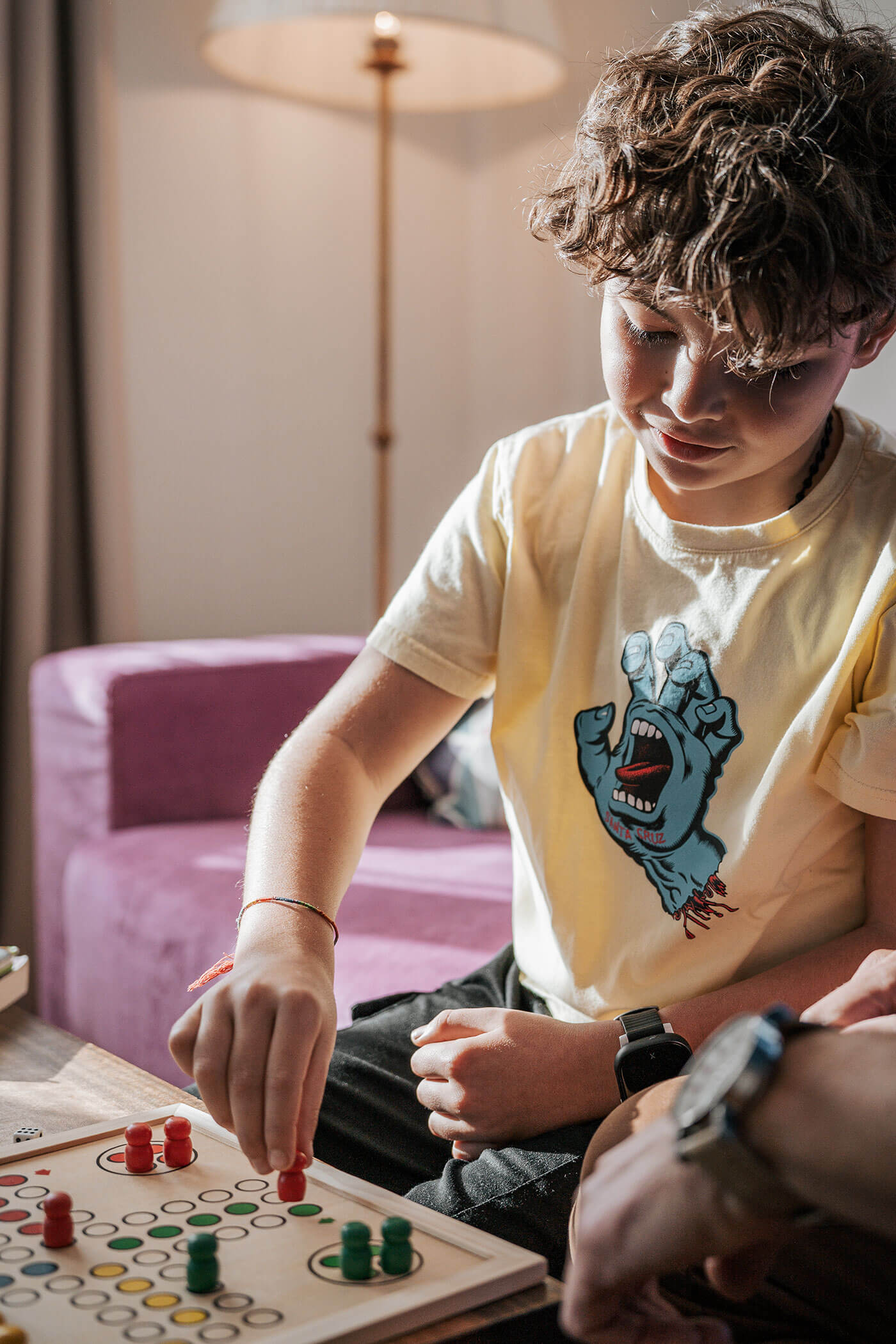 Ein Junge spielt ein Brettspiel, indem er rote Marker auf das Brett legt, während er neben einem lila Sofa sitzt. - Hotel Fameli