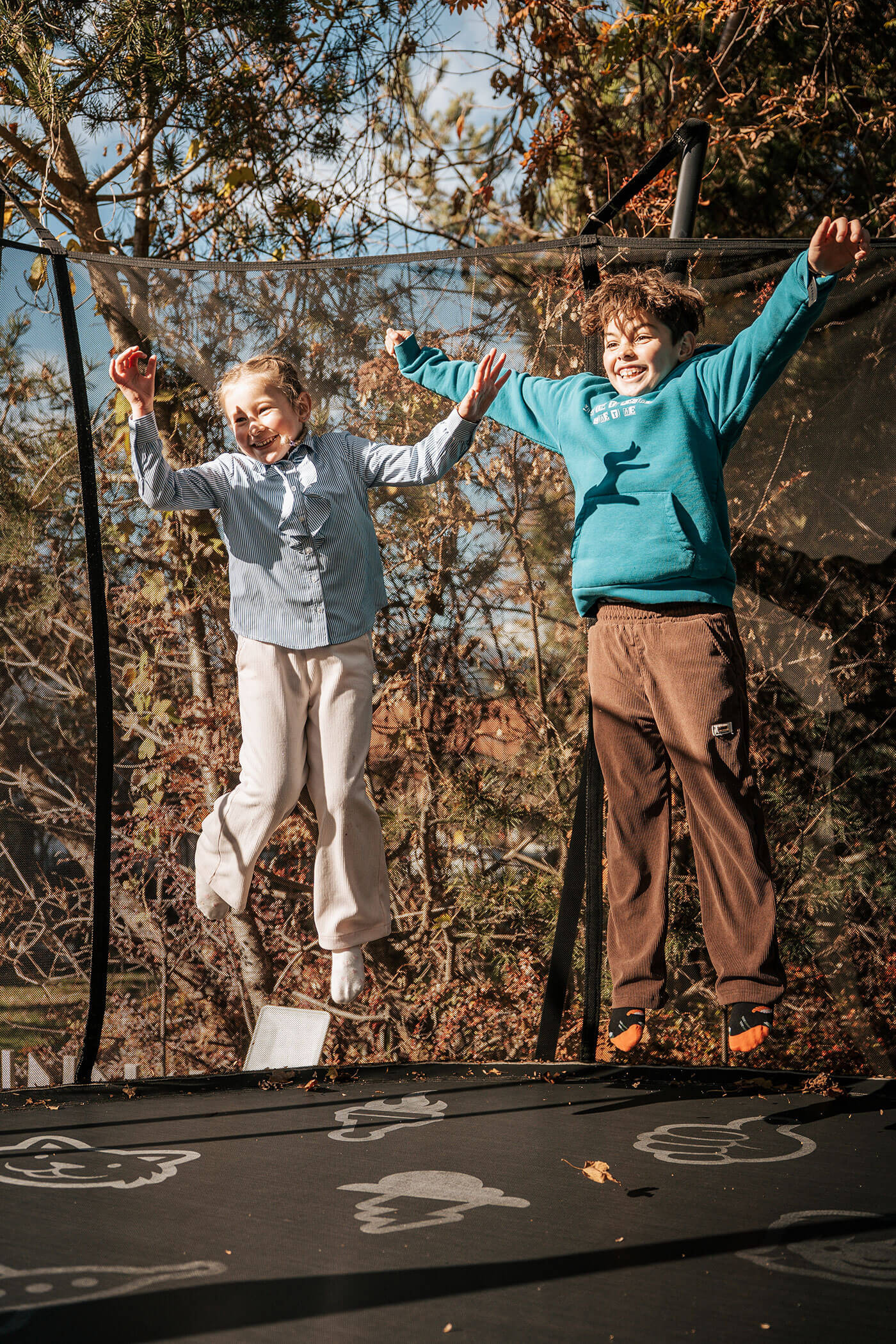 Zwei Kinder springen und lächeln auf einem Trampolin im Freien mit Bäumen im Hintergrund. - Hotel Fameli