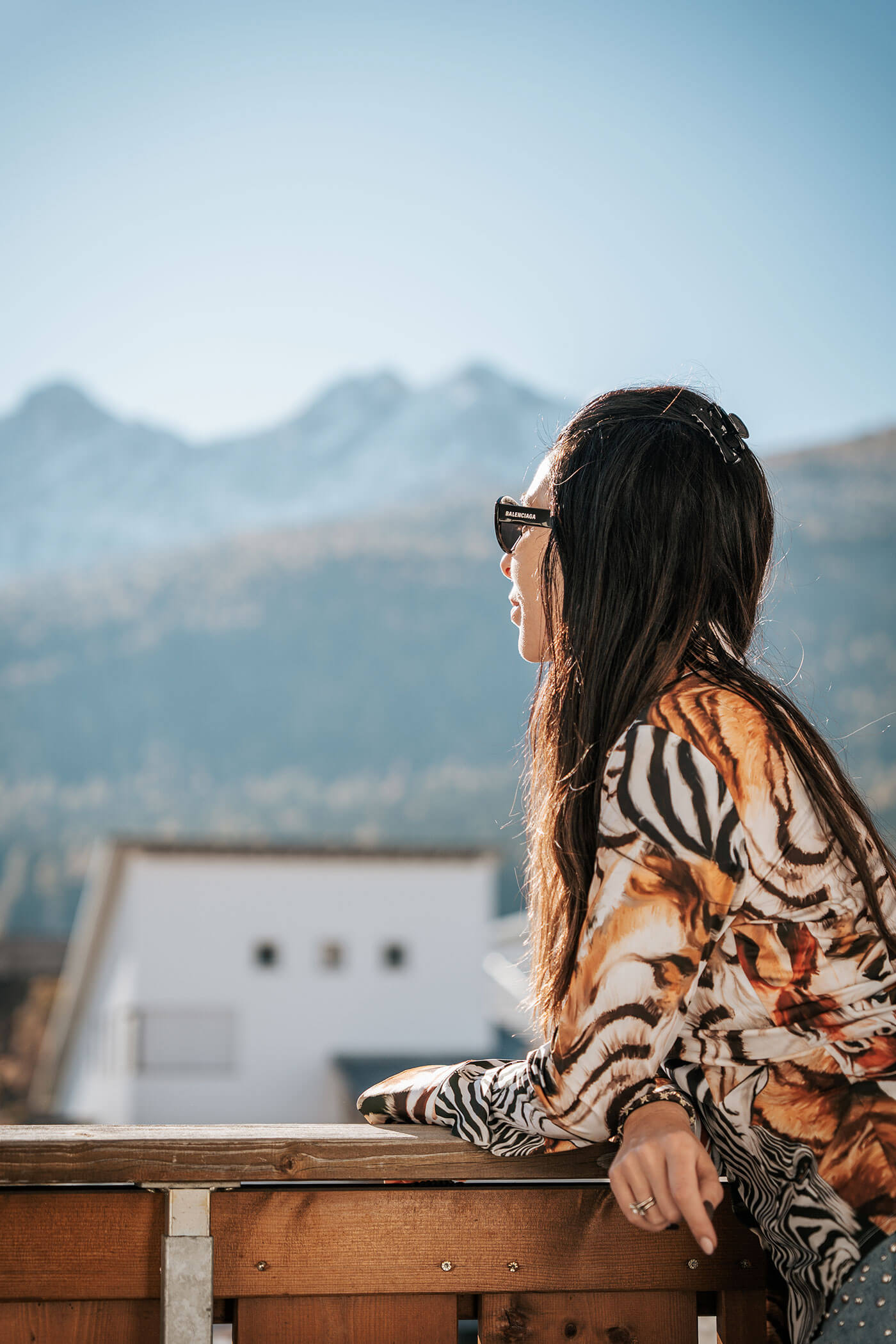 Frau mit Sonnenbrille und langen Haaren blickt an einem sonnigen Tag von einem Holzbalkon auf die fernen Berge. - Hotel Fameli