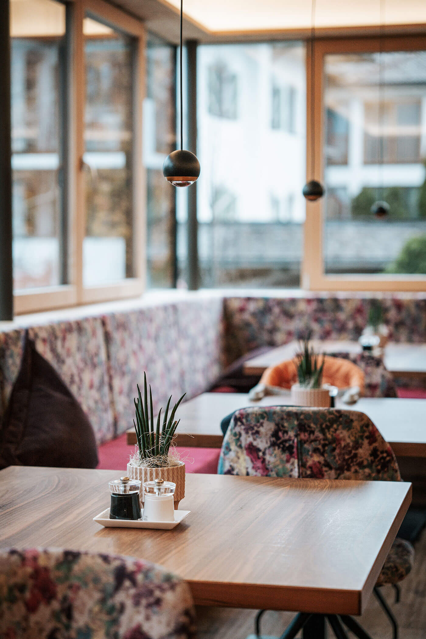 Gemütliches Café-Interieur mit geblümten Stühlen, Holztischen, Topfpflanzen und Hängelampen an großen Fenstern. - Hotel Fameli