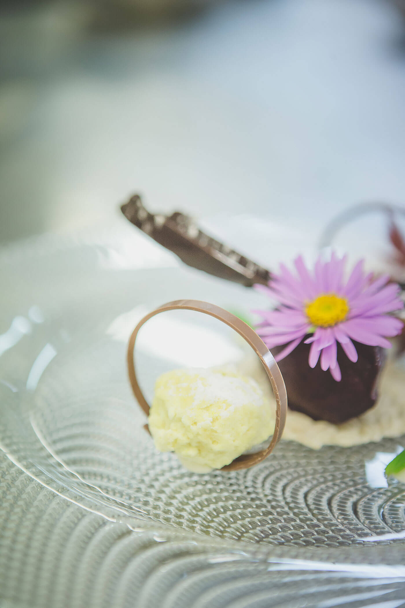 Elegantes Dessert mit Schokolade, einer Kugel Sahne und einer lilafarbenen Blumengarnitur auf einem Teller aus Klarglas. - Hotel Fameli