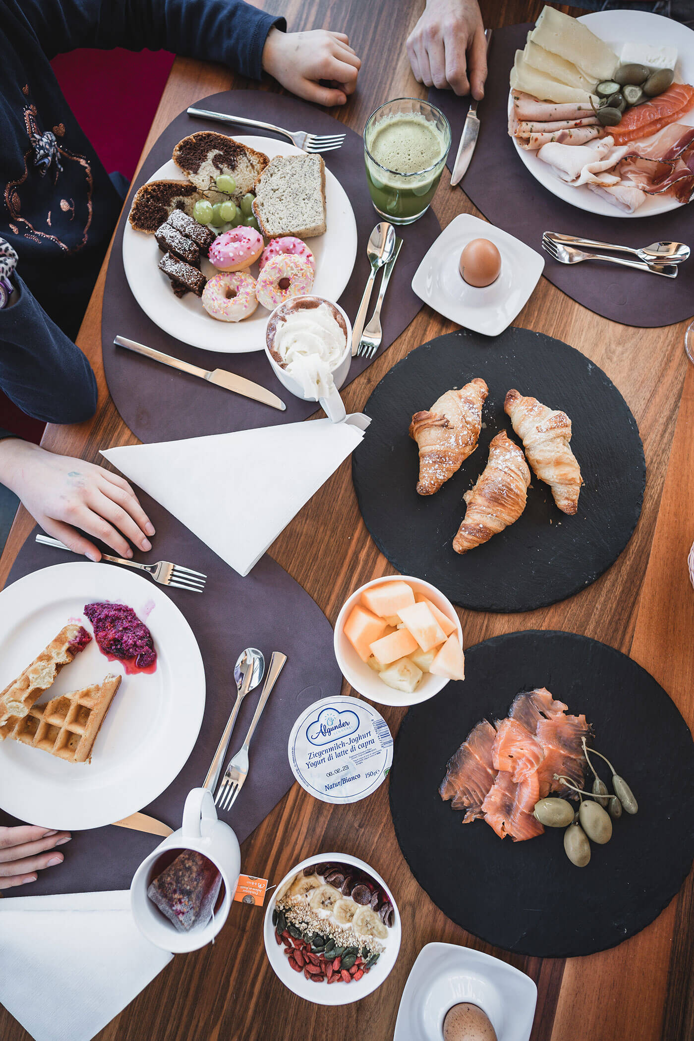 Frühstückstisch mit Croissants, Waffeln, Räucherlachs, Obst, Gebäck und zwei Personen, die nach dem Essen greifen. - Hotel Fameli