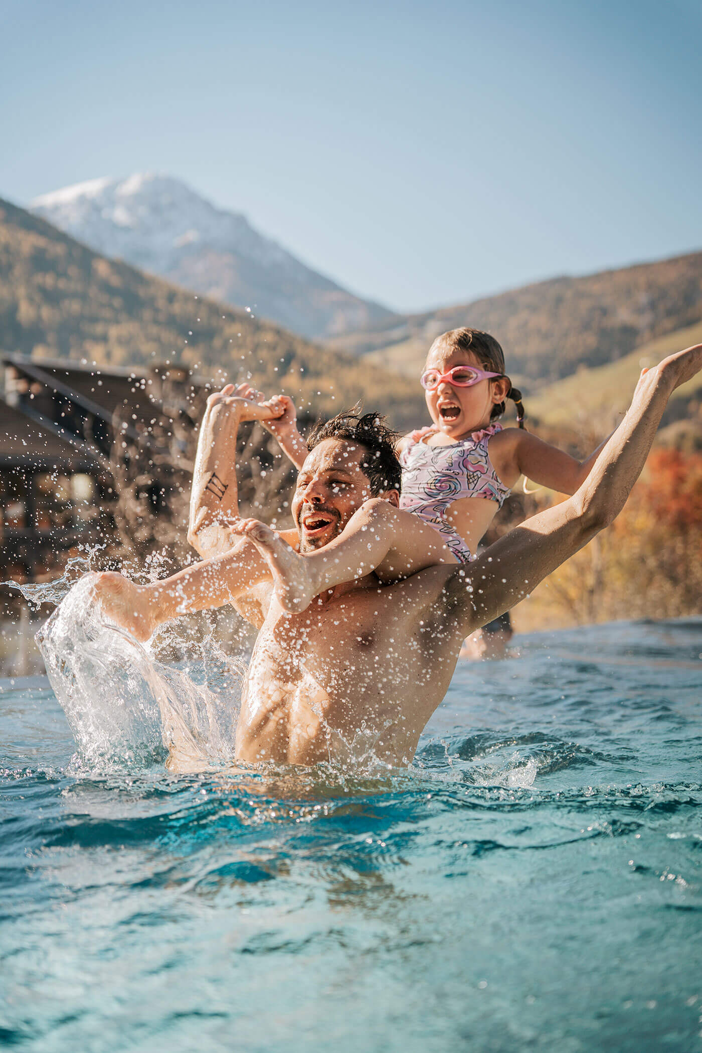 Mann hebt lachendes Mädchen in einen Pool, mit Bergen und Herbstbäumen im Hintergrund an einem sonnigen Tag. - Hotel Fameli