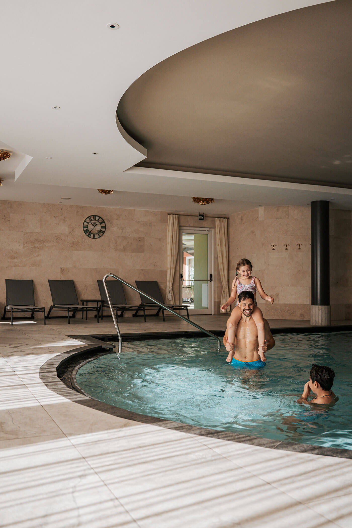 Ein Mann mit einem Mädchen auf den Schultern und ein Junge spielen in einem Hallenbad mit Liegestühlen in der Nähe. - Hotel Fameli
