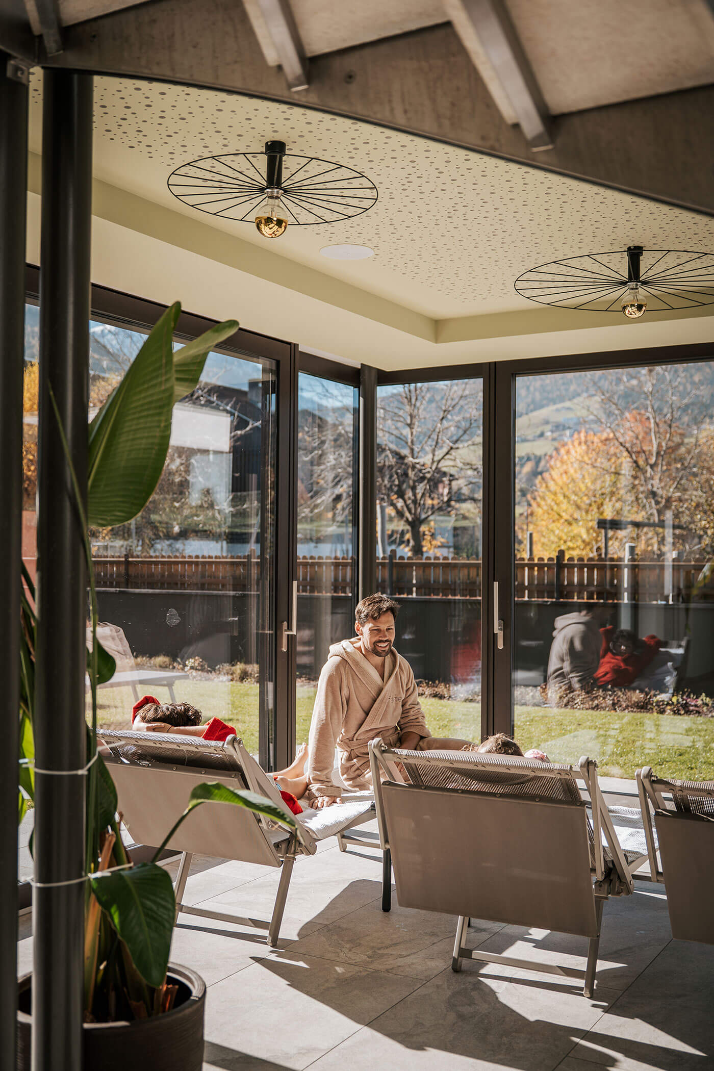 Ein Mann im Bademantel sitzt auf einem Liegestuhl in einem sonnendurchfluteten Raum mit großen Fenstern und Blick auf den Garten. - Hotel Fameli