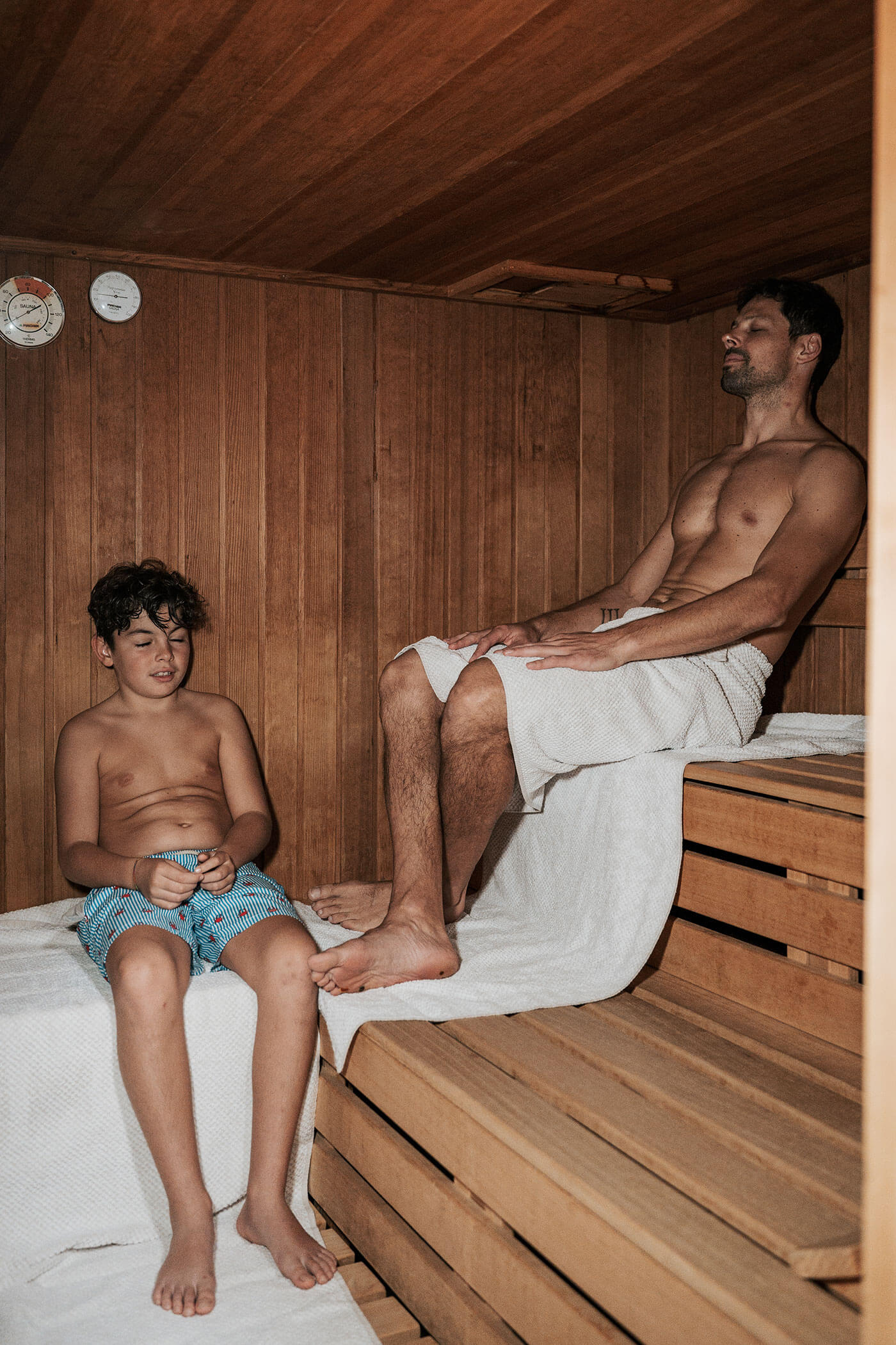 Ein Mann und ein Junge entspannen sich auf Holzbänken in einer Sauna, beide tragen Handtücher um ihre Hüften. - Hotel Fameli
