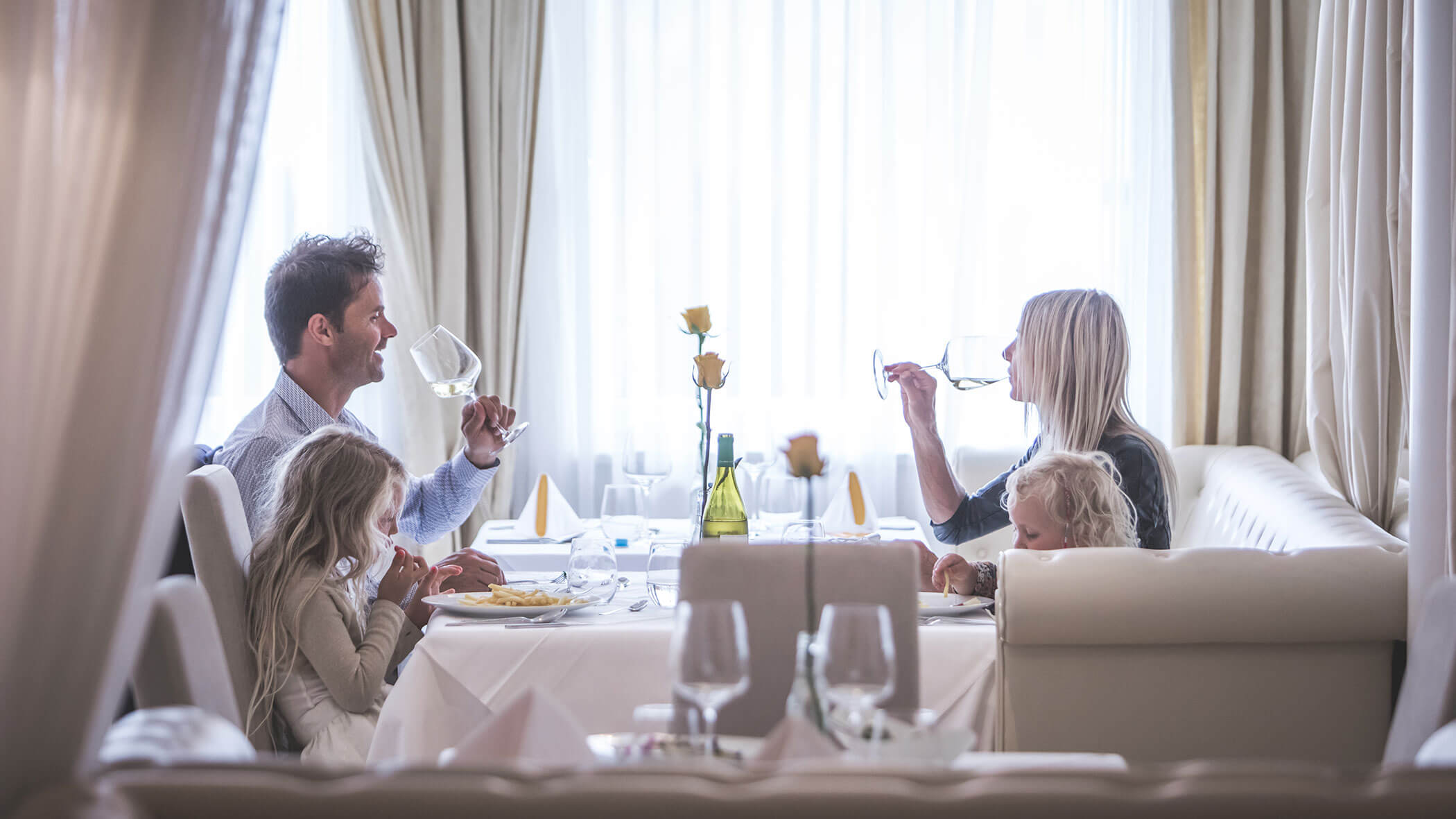 Eine vierköpfige Familie genießt eine gemeinsame Mahlzeit in einem Restaurant mit weißen Tischdecken und gelben Blumen. - Hotel Fameli