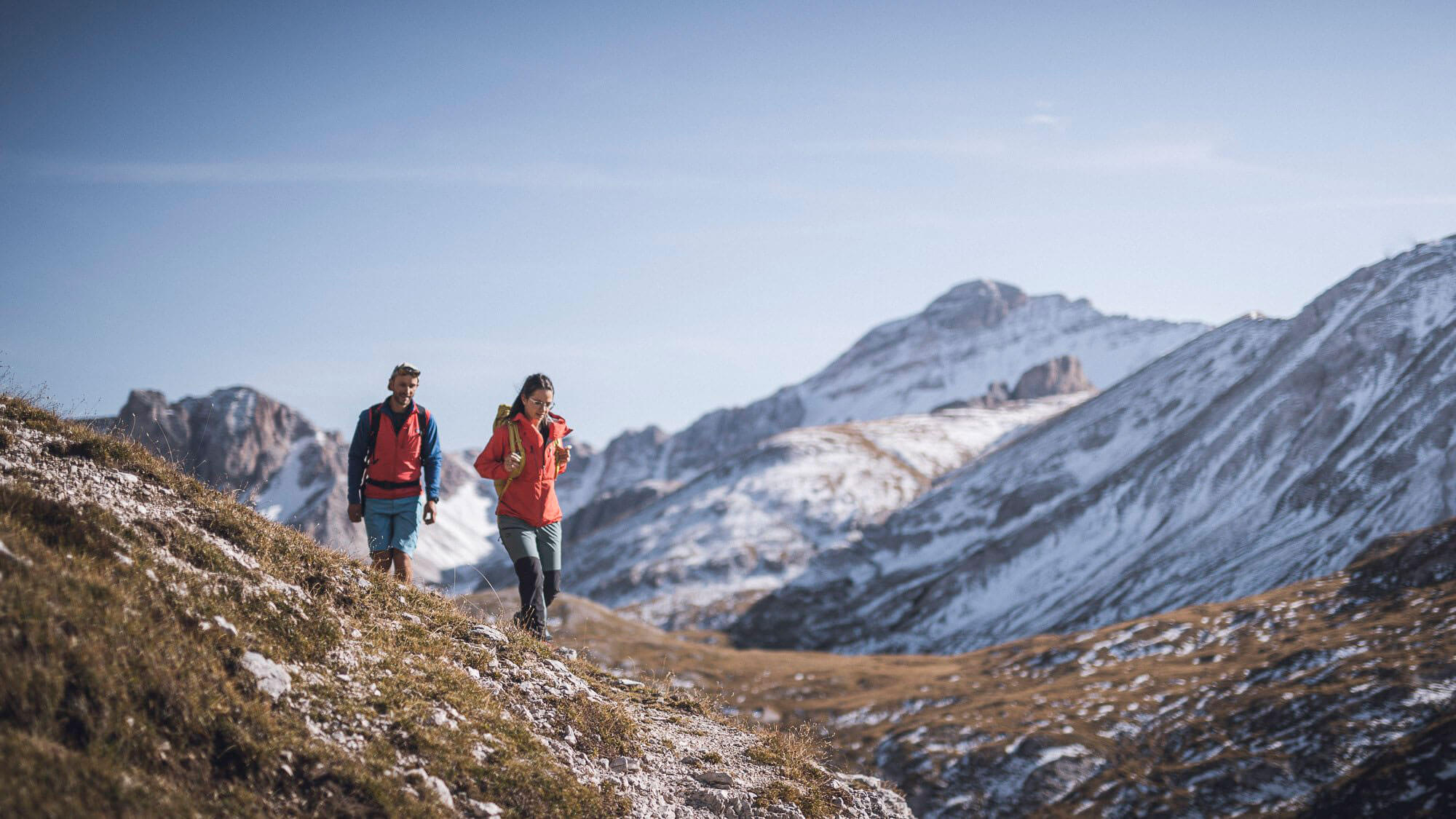 Zwei Wanderer mit Rucksäcken gehen auf einem Bergpfad mit schneebedeckten Gipfeln im Hintergrund unter einem klaren Himmel. - Hotel Fameli