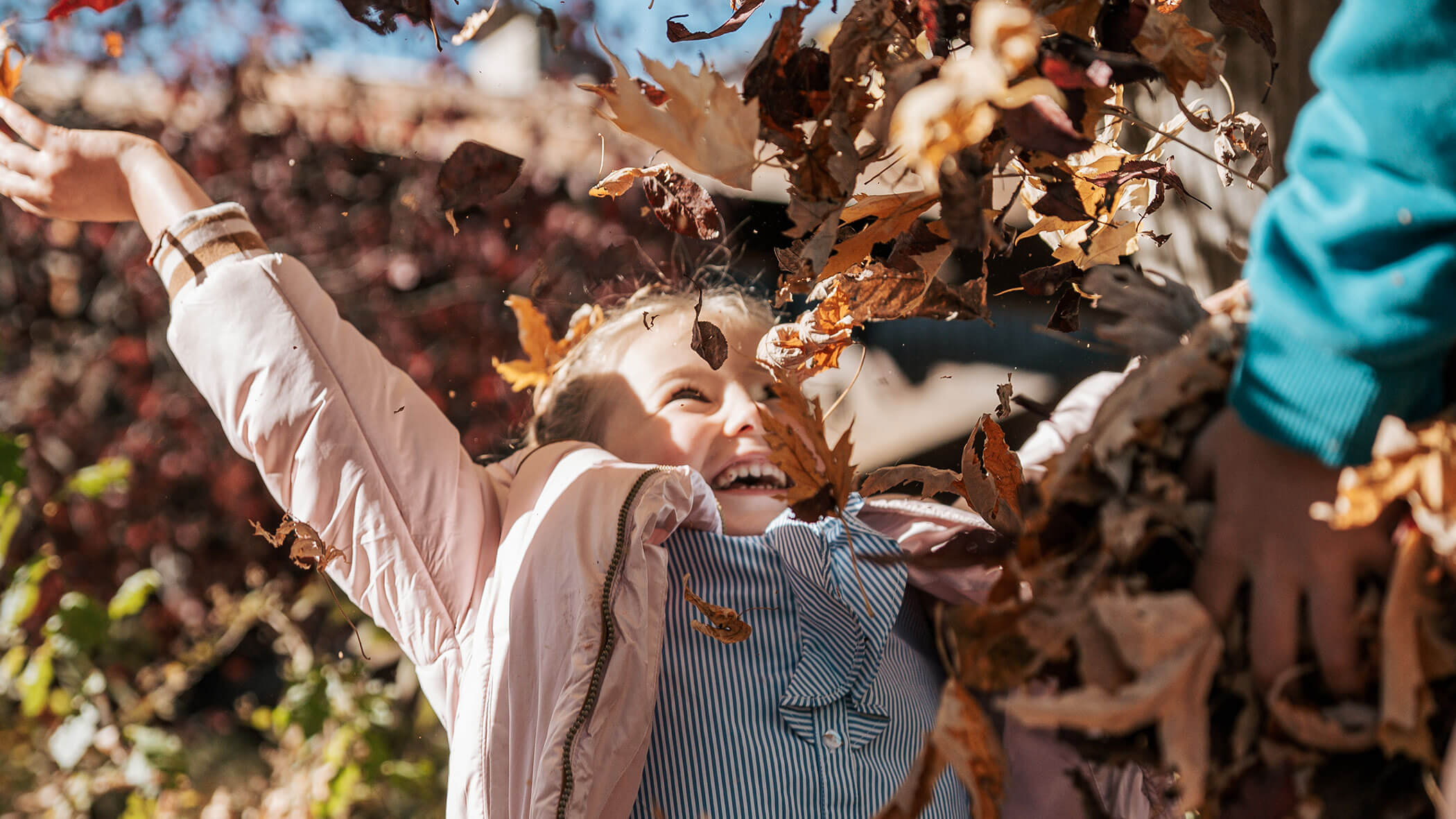 Ein lächelndes Kind in einer Jacke wirft Herbstblätter in die Luft und genießt einen sonnigen Herbsttag im Freien. - Hotel Fameli