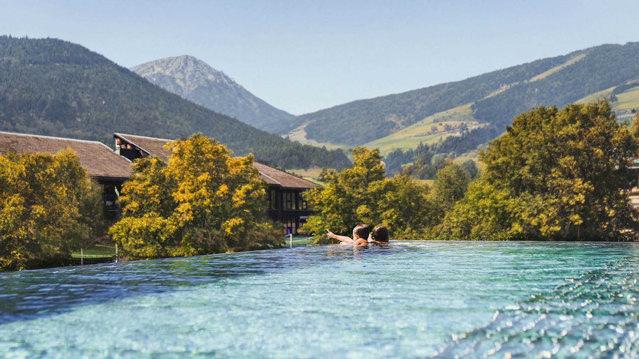 Eine Person entspannt sich in einem Infinity-Pool im Freien mit grünen Hügeln und Bergen im Hintergrund. - Hotel Fameli