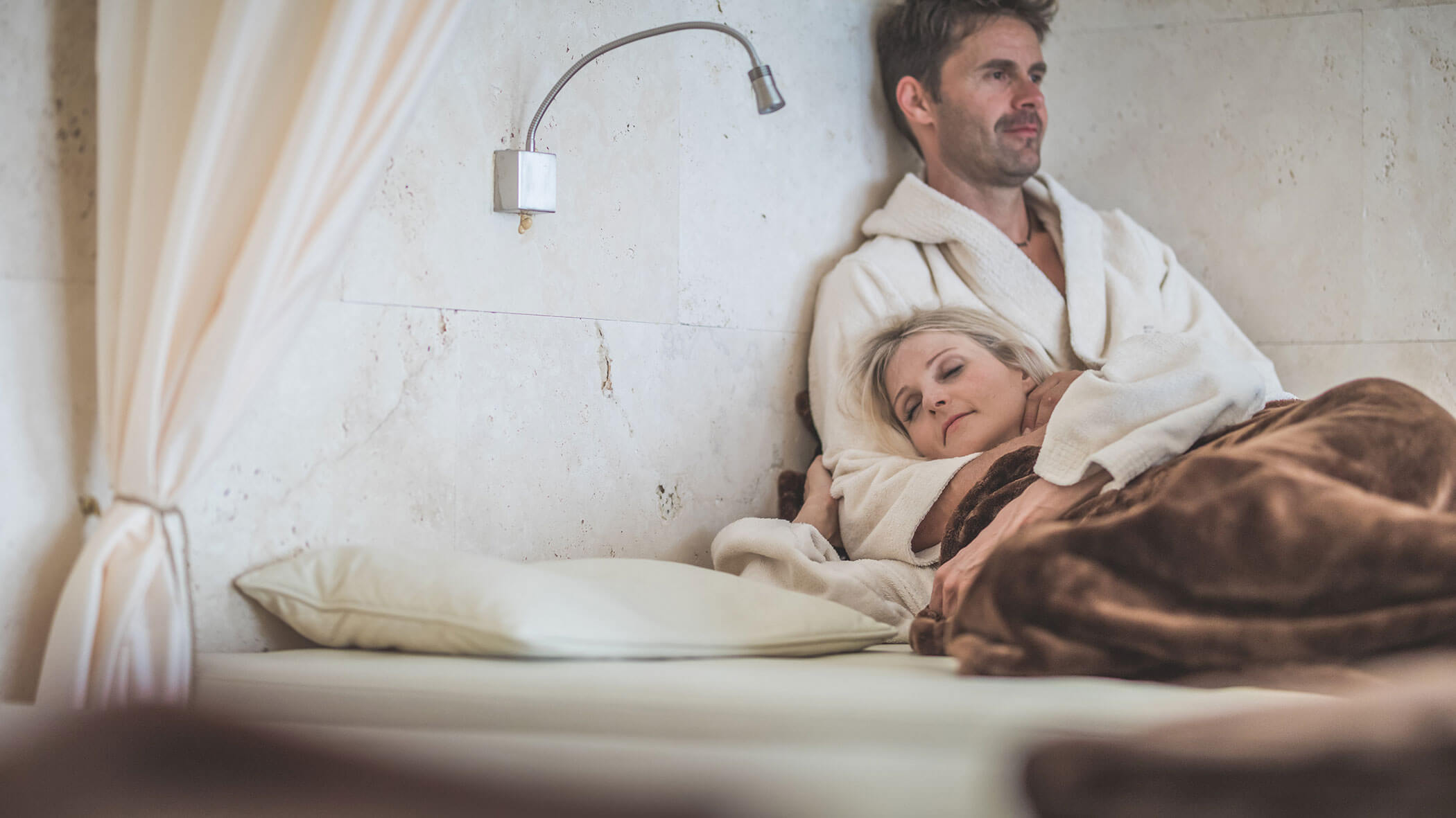 Ein Paar in Bademänteln entspannt sich im Bett, wobei die Frau ihren Kopf unter einer braunen Decke auf der Brust des Mannes ruht. - Hotel Fameli