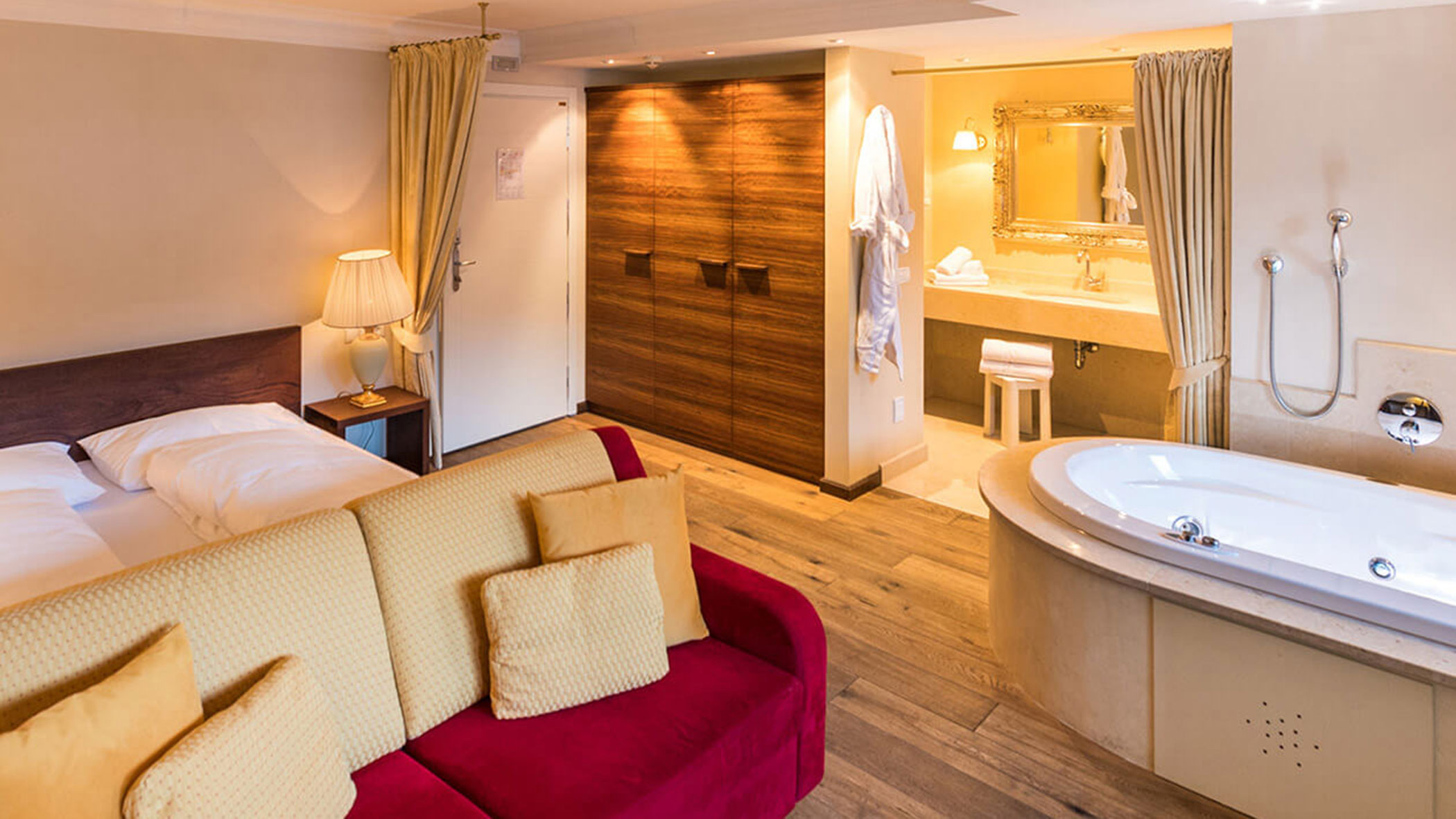Hotelzimmer mit Doppelbett, rotem Sofa, offenem Schrank, Bademantel und Badewanne in der Nähe des Waschbeckens. - Hotel Fameli