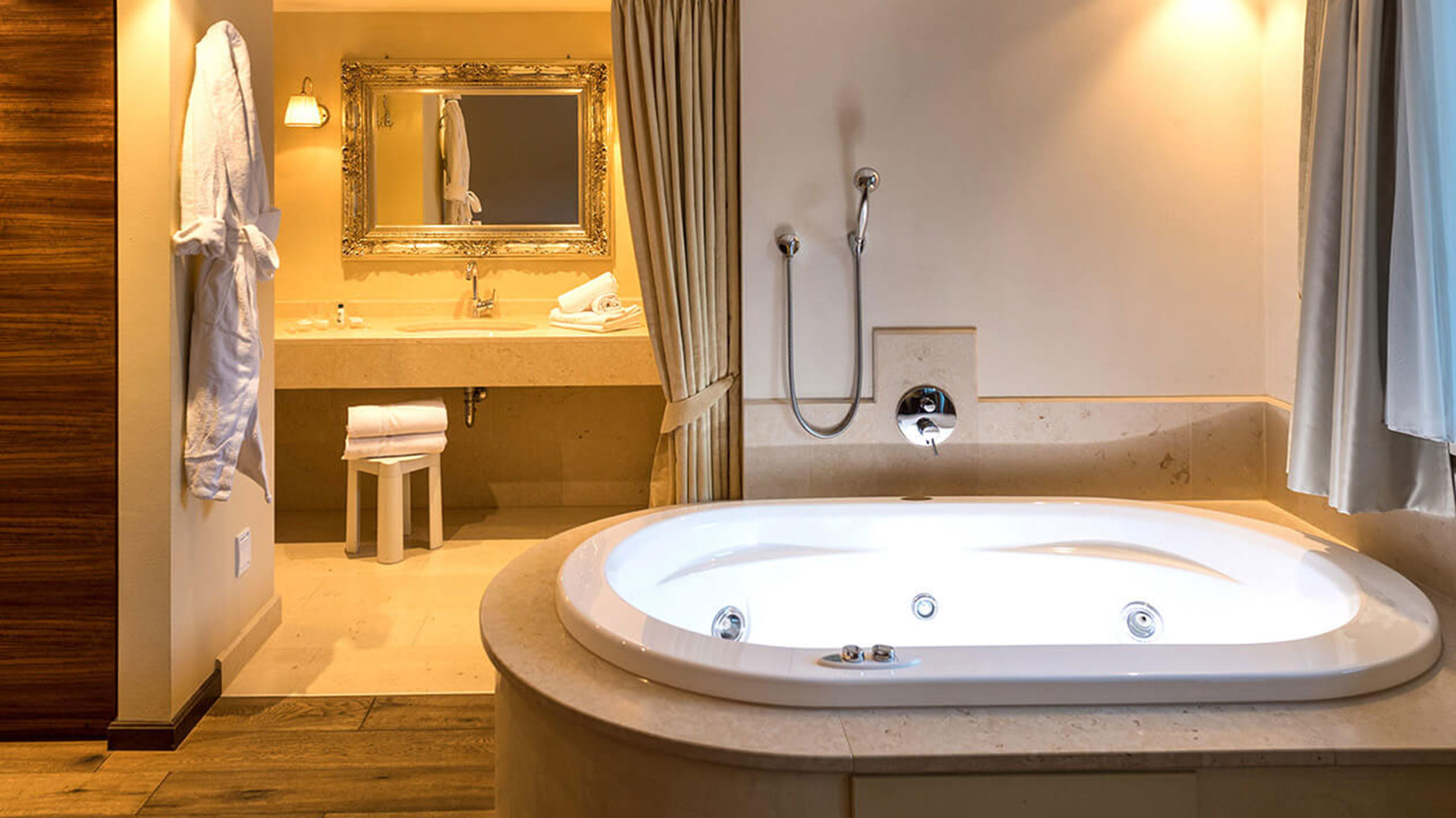 Große Badewanne mit weißen Handtüchern, Bademantel und Spiegel in einem warm beleuchteten, eleganten Badezimmer. - Hotel Fameli