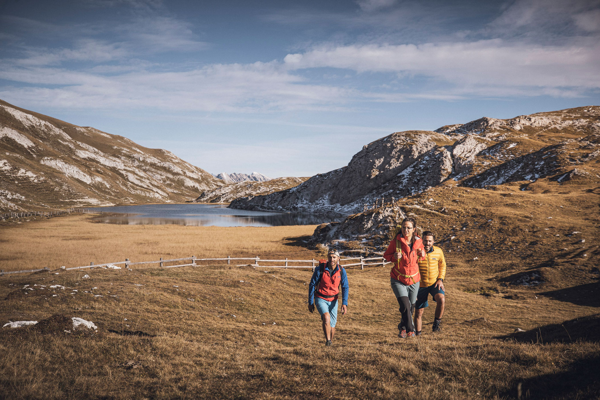 Drei Personen wandern auf einen grasbewachsenen Hügel mit Bergen und einem See im Hintergrund unter einem blauen Himmel. - Hotel Fameli