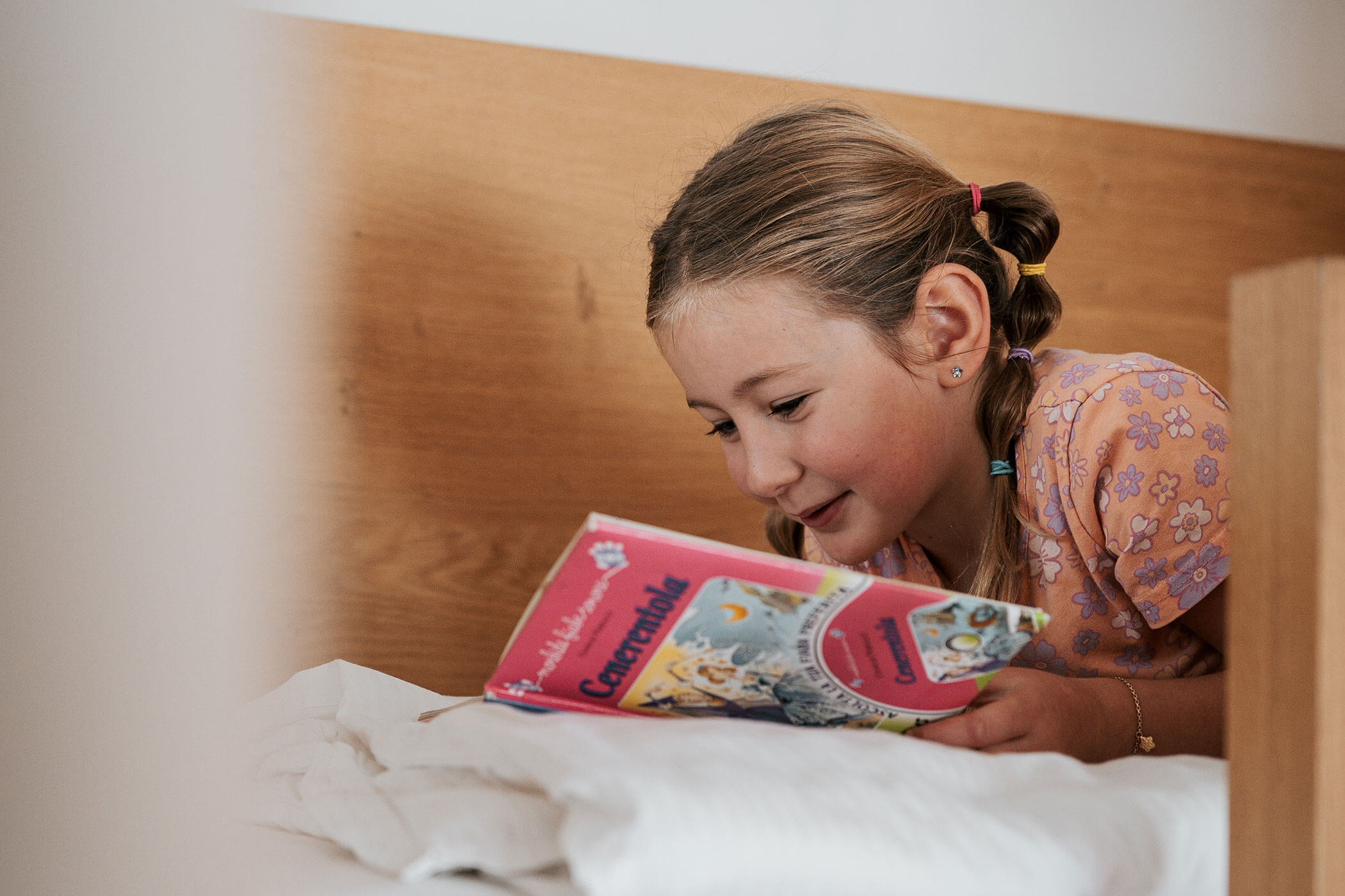 Ein junges Mädchen liegt lächelnd auf einem Bett und liest ein buntes Kinderbuch. - Hotel Fameli