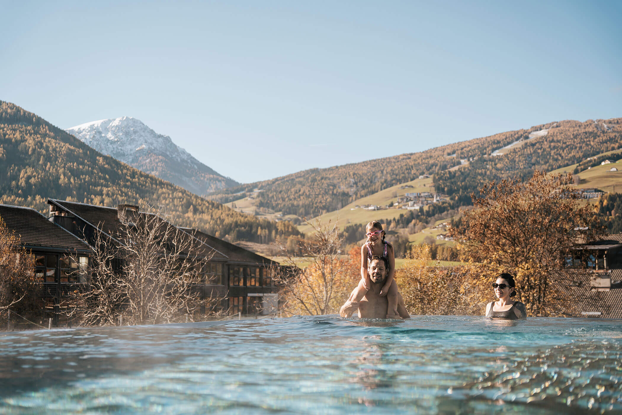 Drei Personen genießen einen Außenpool mit Bergen und herbstlichen Bäumen im Hintergrund unter einem klaren Himmel. - Hotel Fameli