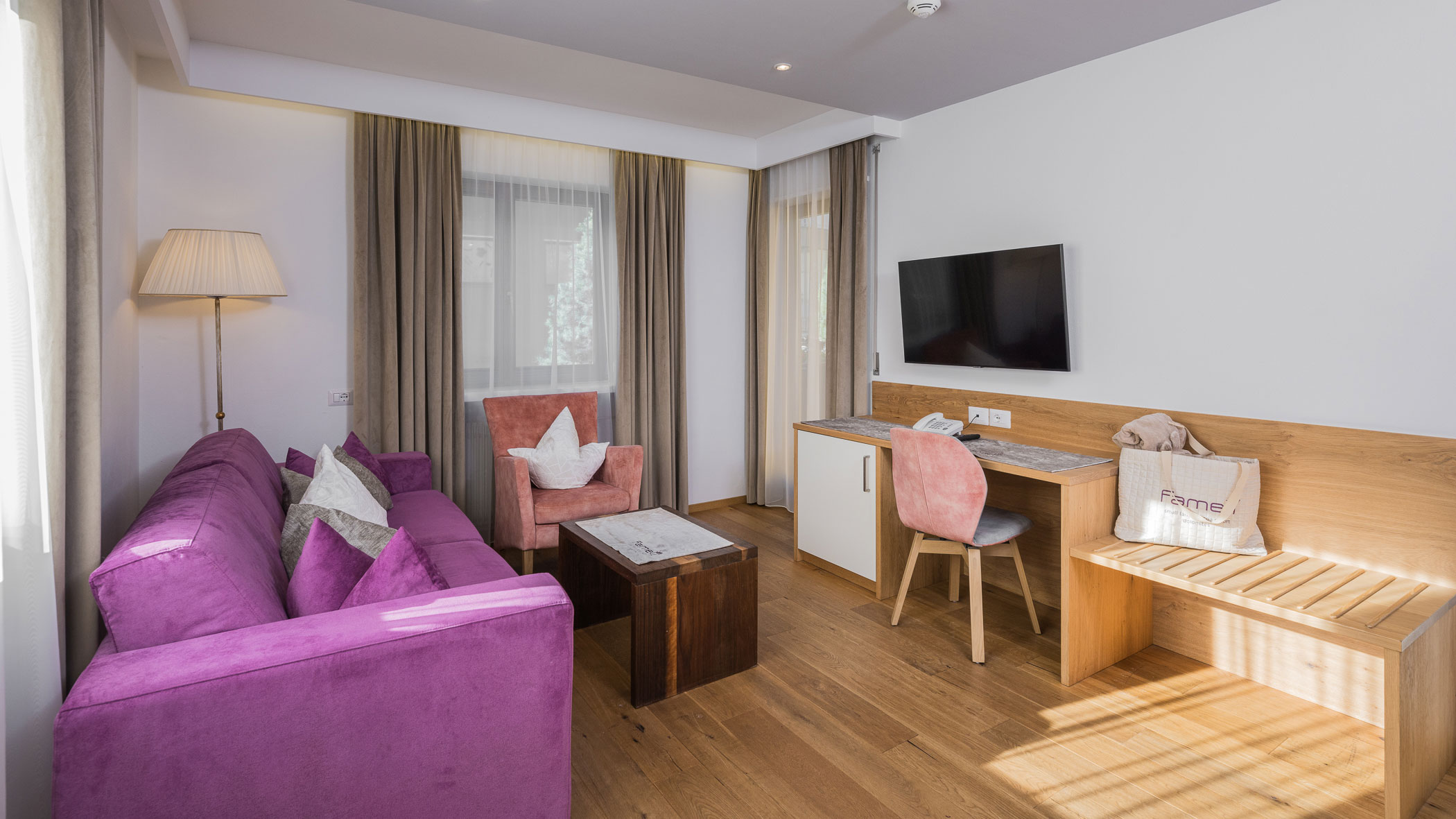 Modernes Wohnzimmer mit lilafarbenem Sofa, rosafarbenem Sessel, Fernseher, Schreibtisch und natürlichem Licht durch große Fenster. - Hotel Fameli