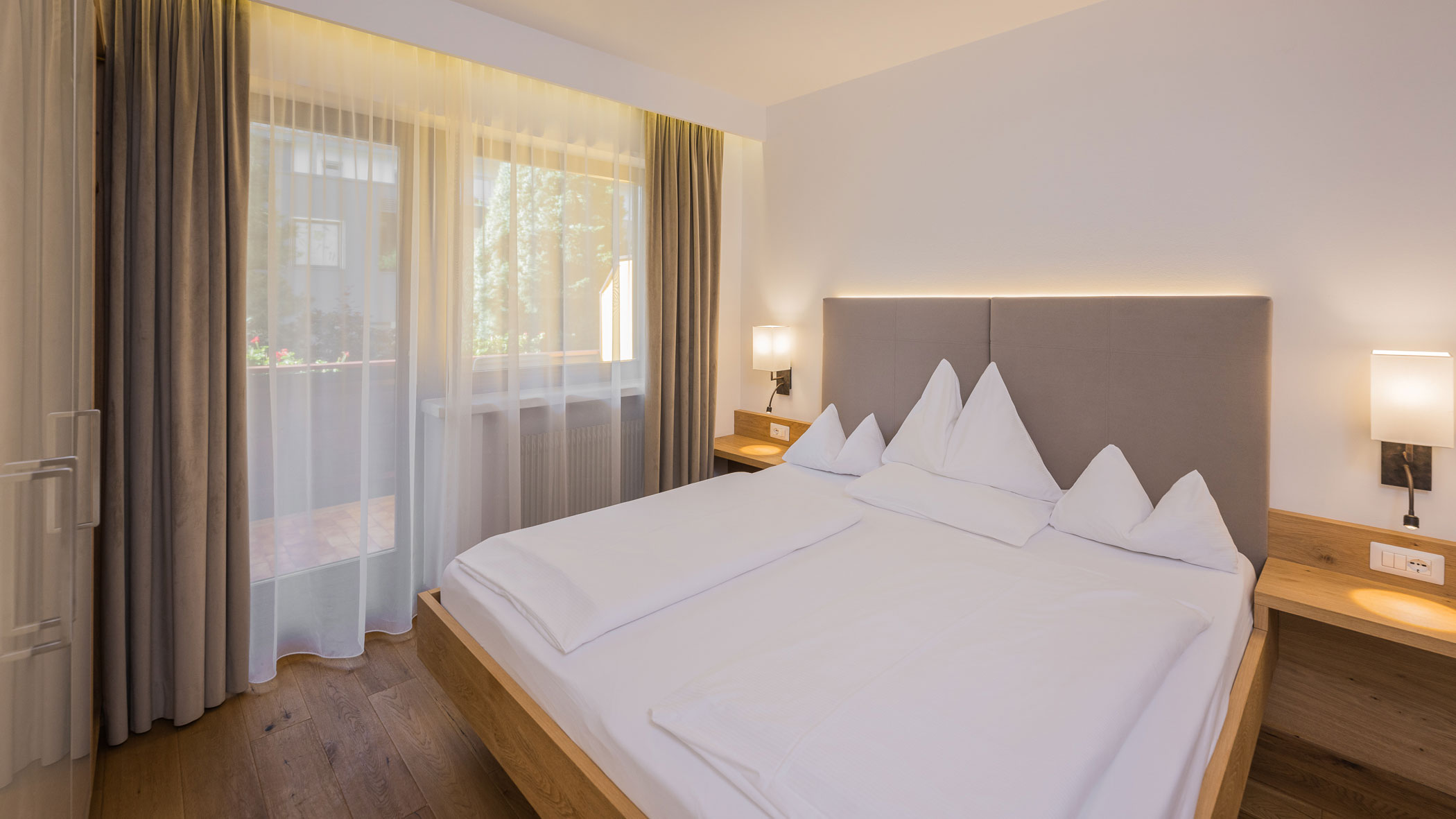 Modernes Hotelzimmer mit Doppelbett, weißer Bettwäsche, Nachttischen und einem großen Fenster mit durchsichtigen Vorhängen. - Hotel Fameli