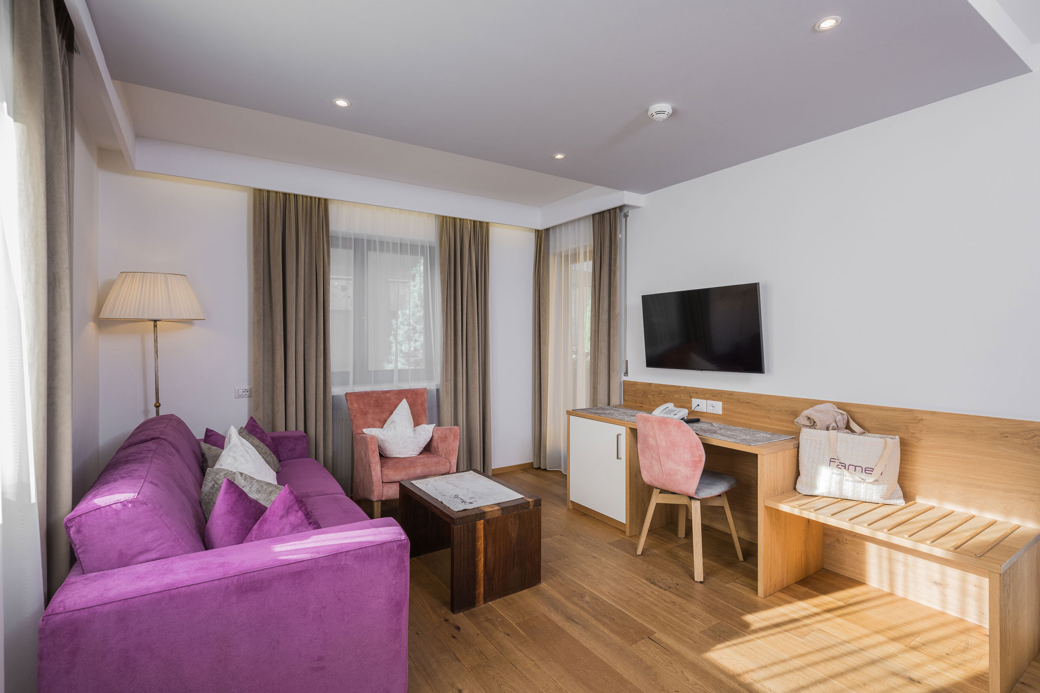 Helles Wohnzimmer mit lilafarbenem Sofa, Schreibtisch, TV und Holzfußboden, beleuchtet von Tages- und Deckenlicht. - Hotel Fameli