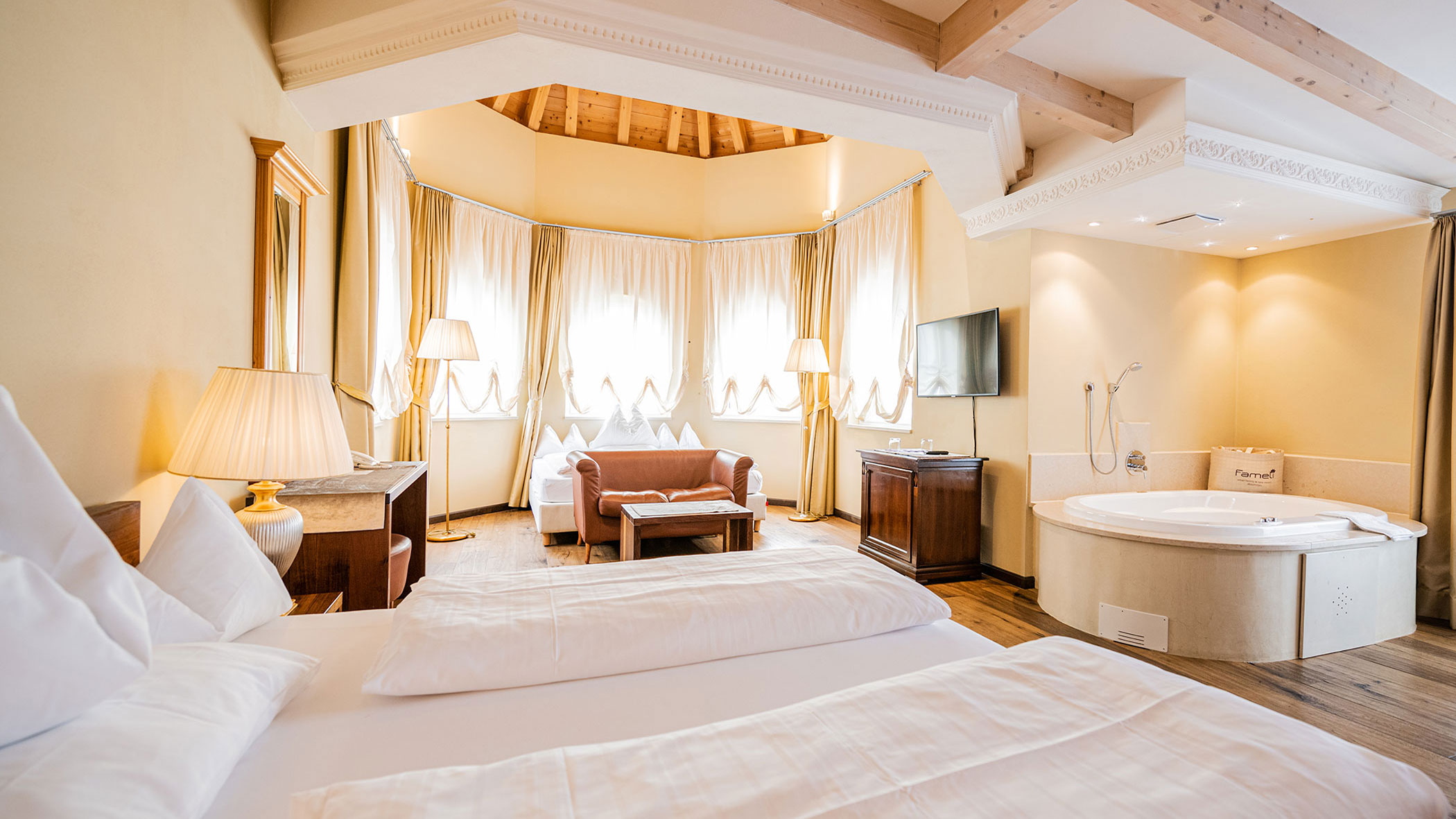Helles Hotelzimmer mit einem großen Bett, Sofa, Fernseher, Lampen und einer runden Badewanne in der Ecke. - Hotel Fameli