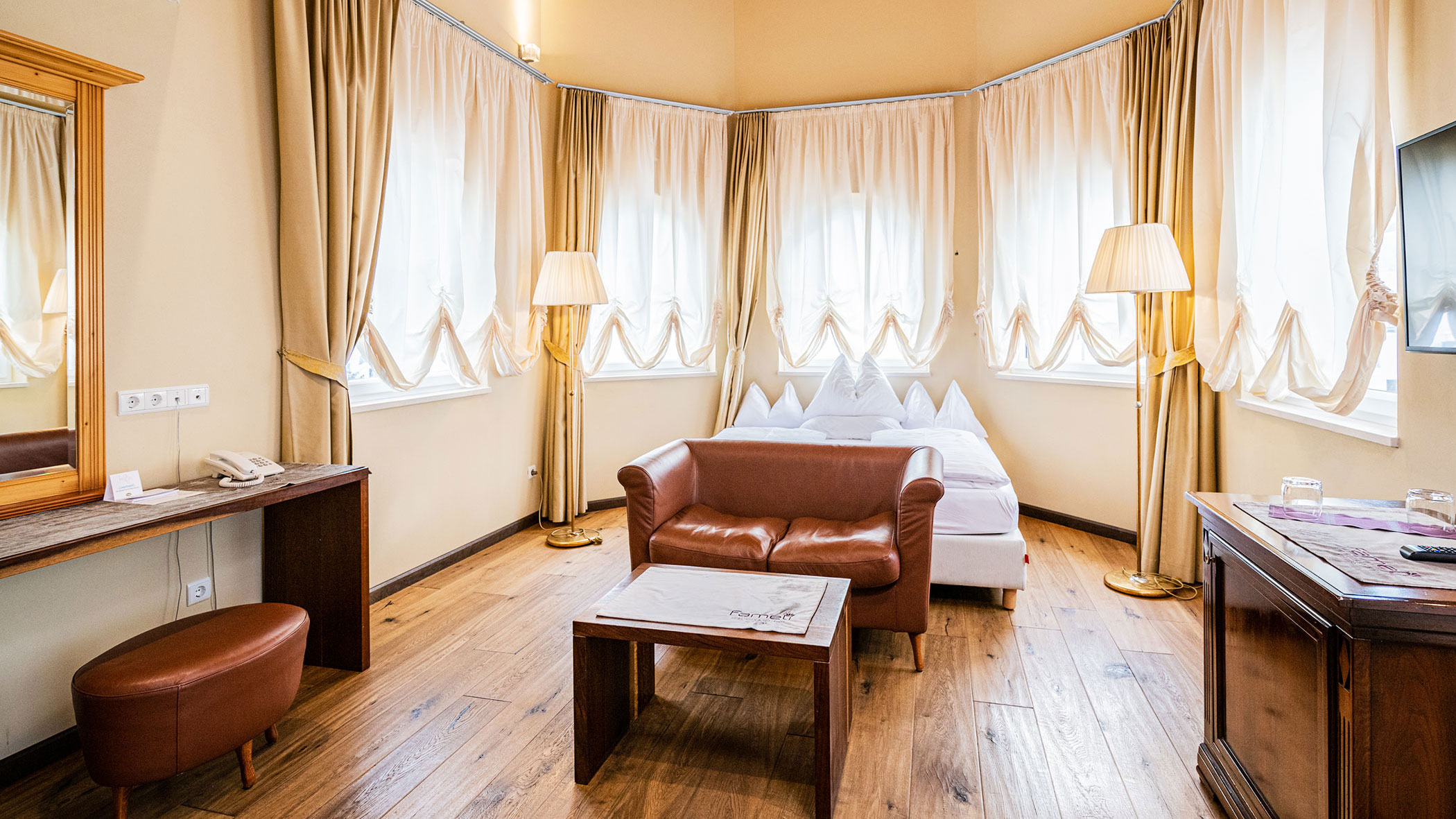 Ein helles Hotelzimmer mit einem Bett, einem braunen Ledersofa, einem Schreibtisch, Lampen und großen Fenstern mit cremefarbenen Vorhängen. - Hotel Fameli