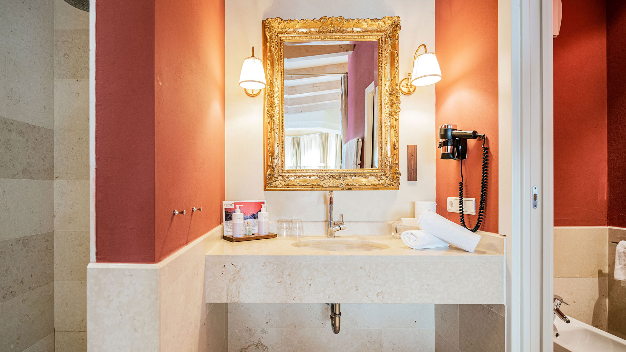 Elegantes Badezimmer mit goldgerahmtem Spiegel, Waschbecken, gerollten Handtüchern und Wandleuchten vor roten und cremefarbenen Wänden. - Hotel Fameli