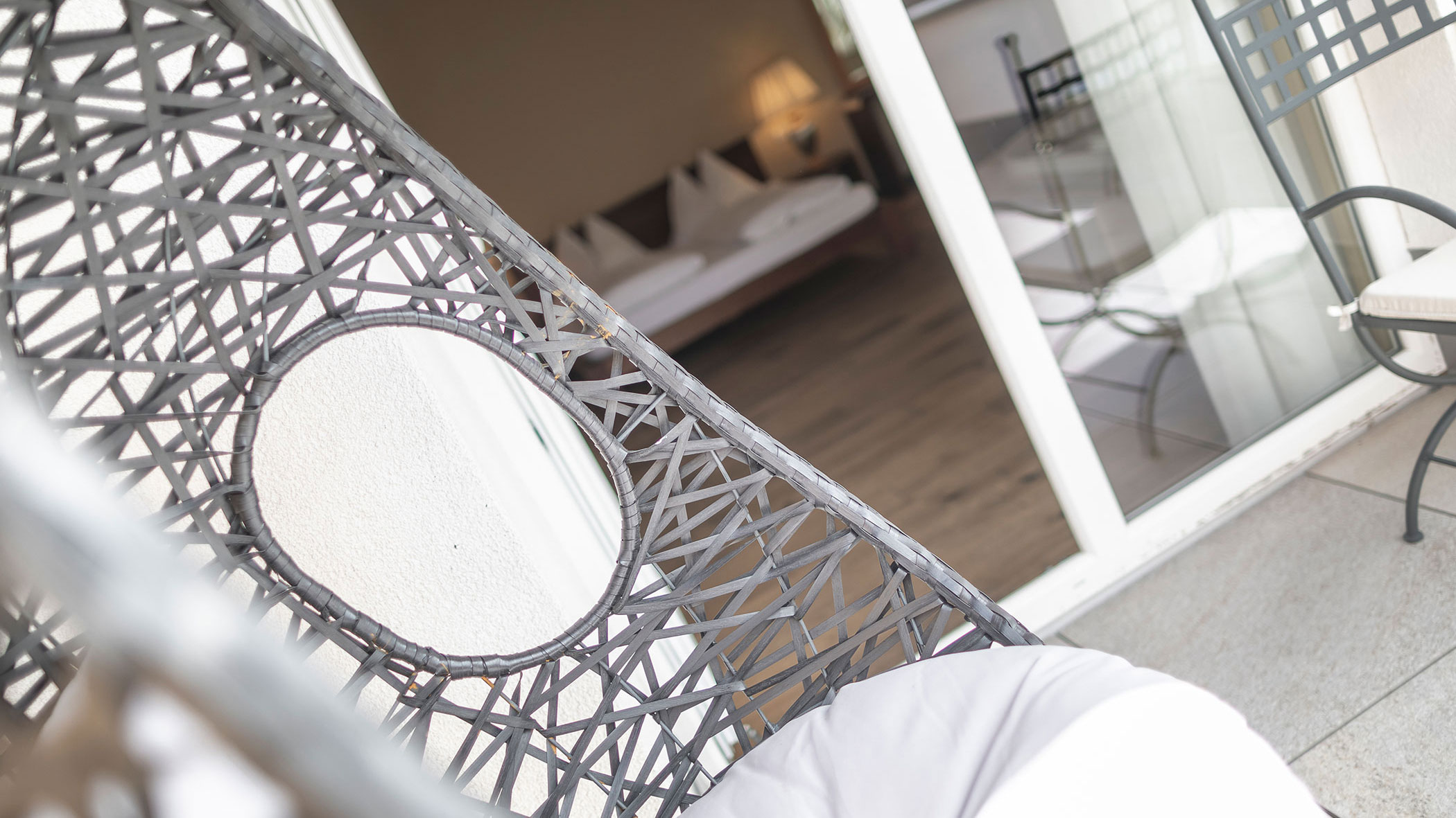 Hängesessel aus Korbgeflecht auf einem Balkon mit Blick in ein Schlafzimmer mit einem gemachten Bett und einer Lampe. - Hotel Fameli