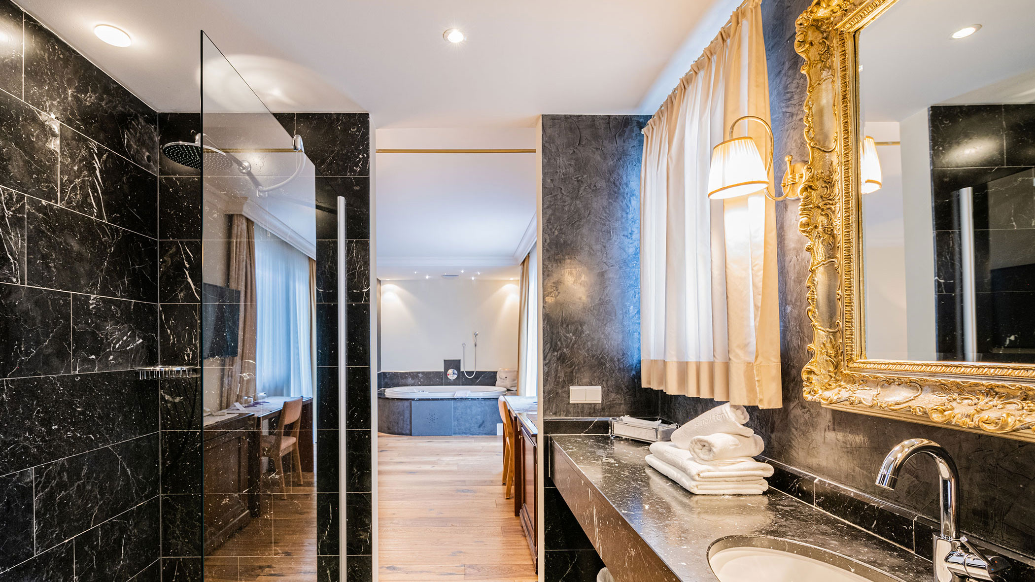 Luxuriöses, modernes Badezimmer mit schwarzem Marmor, goldgerahmtem Spiegel und Blick auf eine Badewanne im Nebenraum. - Hotel Fameli