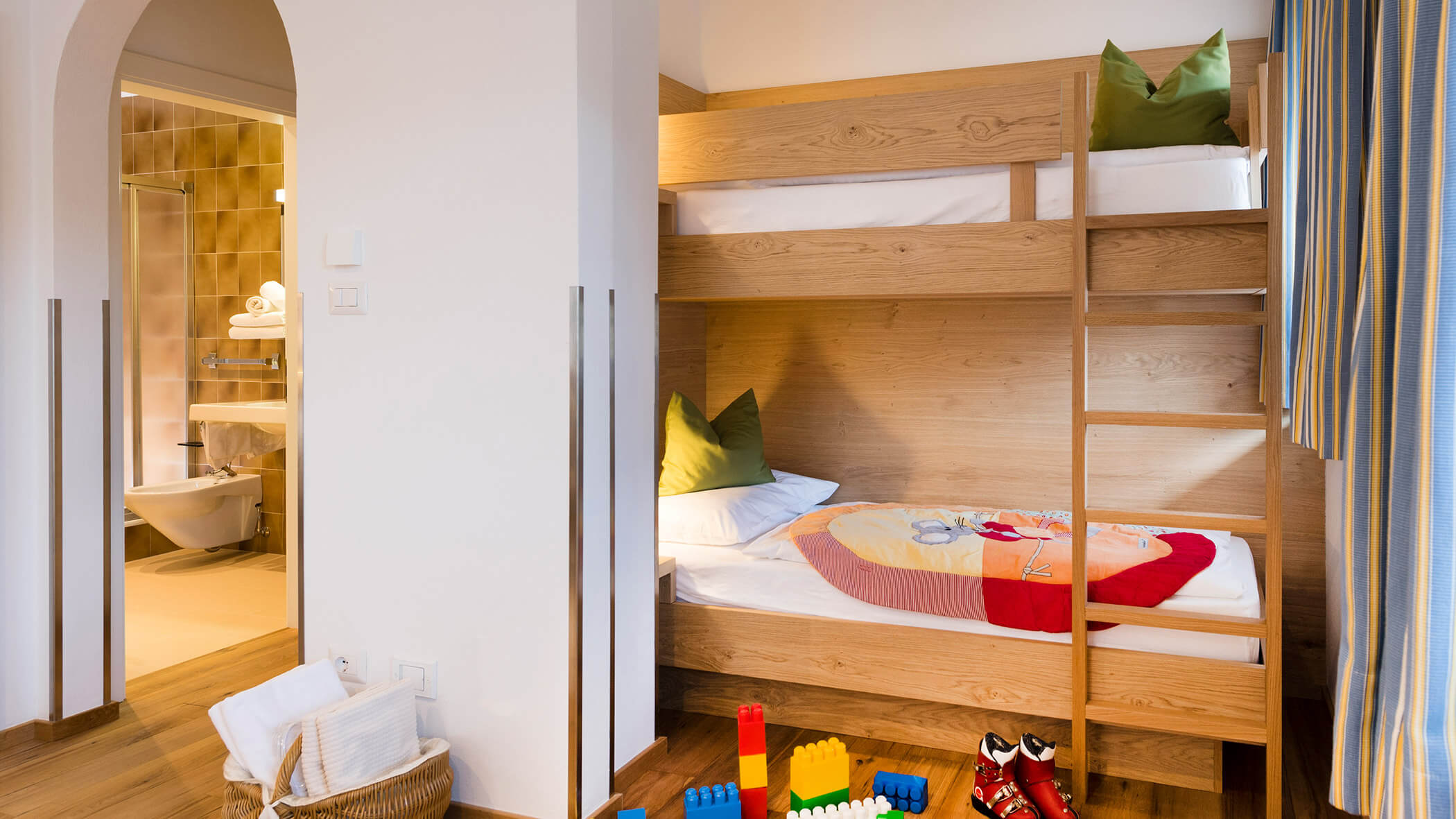 Etagenbetten aus Holz mit grünen Kissen in einem gemütlichen Zimmer, Spielzeug und Schuhe auf dem Boden und ein Badezimmer in Sichtweite. - Hotel Fameli