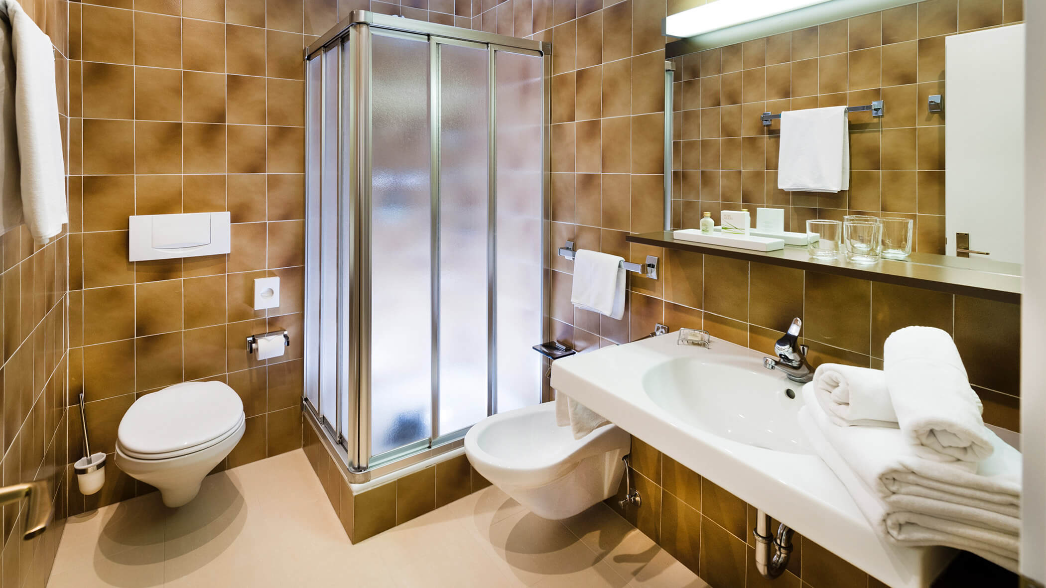 Modernes Badezimmer mit braun gefliesten Wänden, Glasdusche, weißem Waschbecken, Toilette und ordentlich gefalteten Handtüchern. - Hotel Fameli