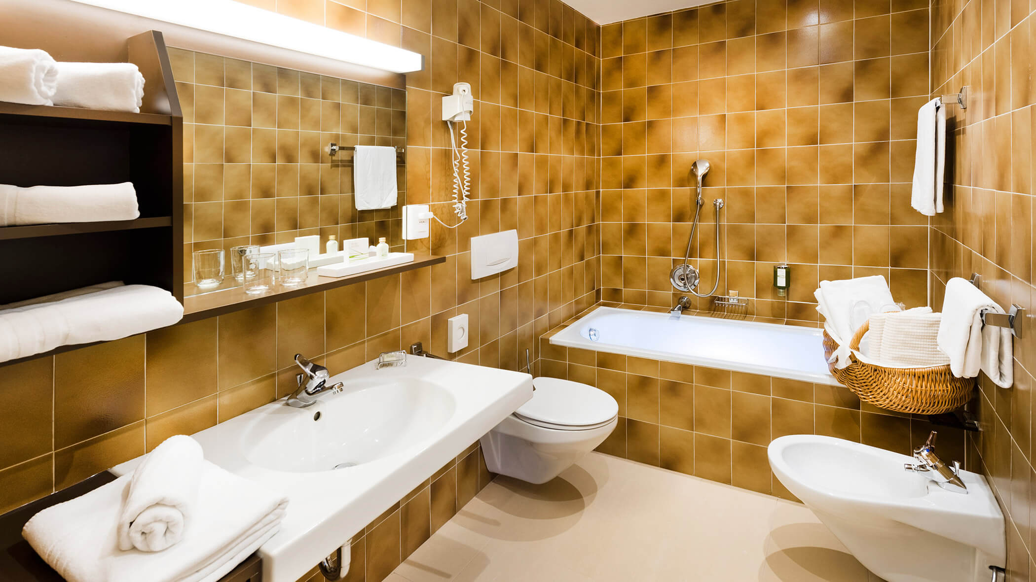 Badezimmer mit braun gefliesten Wänden, weißem Waschbecken, Toilette, Bidet, Badewanne, Handtüchern und Toilettenartikeln in Regalen. - Hotel Fameli