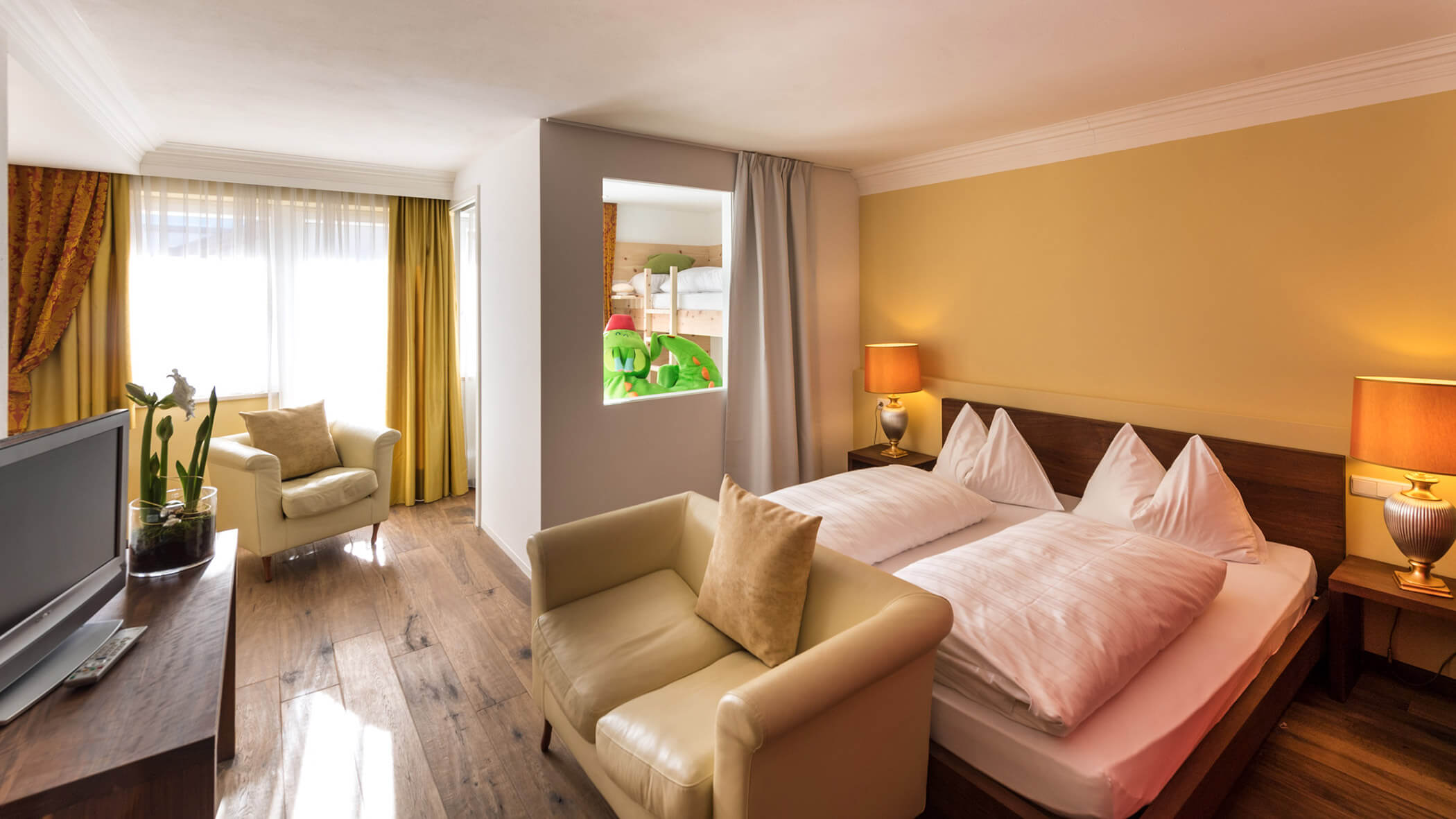 Helles Hotelzimmer mit Doppelbett, Sesseln, TV, Lampen und einer Trennwand, hinter der sich Etagenbetten befinden. - Hotel Fameli