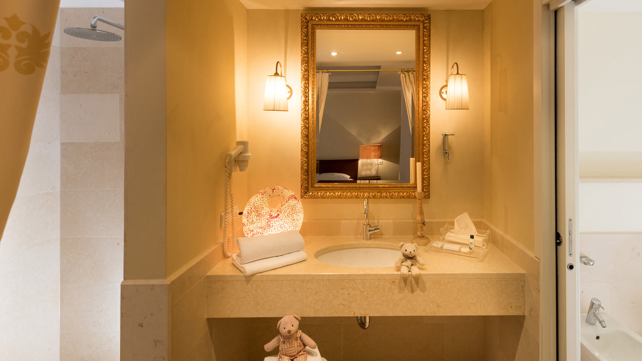Elegantes Badezimmer mit Marmorwaschbecken, goldgerahmtem Spiegel, sanfter Beleuchtung und zwei Plüschteddybären auf dem Tresen. - Hotel Fameli