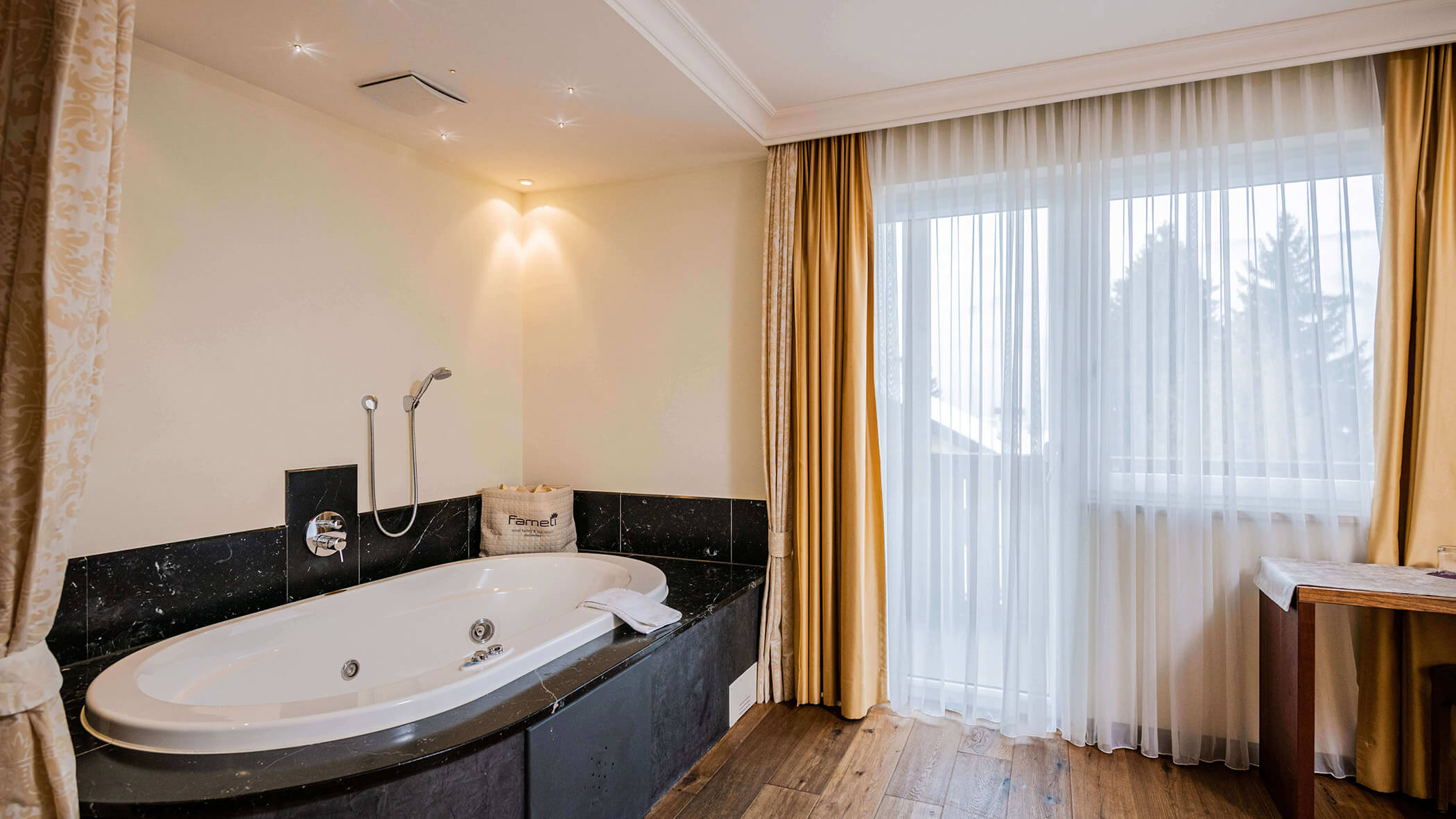 Modernes Badezimmer mit einer großen ovalen Badewanne, schwarzen Marmorakzenten und transparenten Vorhängen, die natürliches Licht hereinlassen. - Hotel Fameli